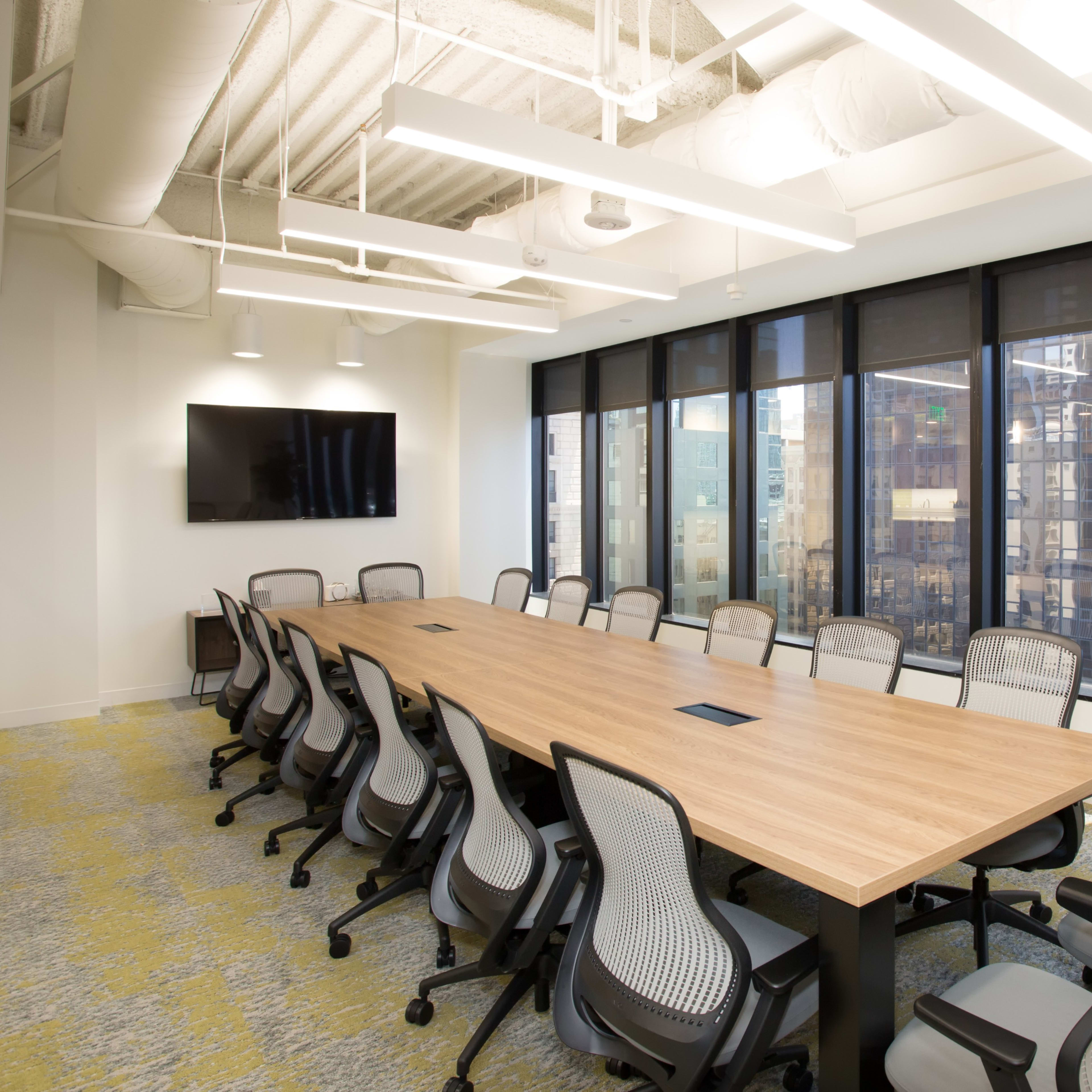 A large conference room features a long wooden table surrounded by ergonomic chairs and large windows with city views.