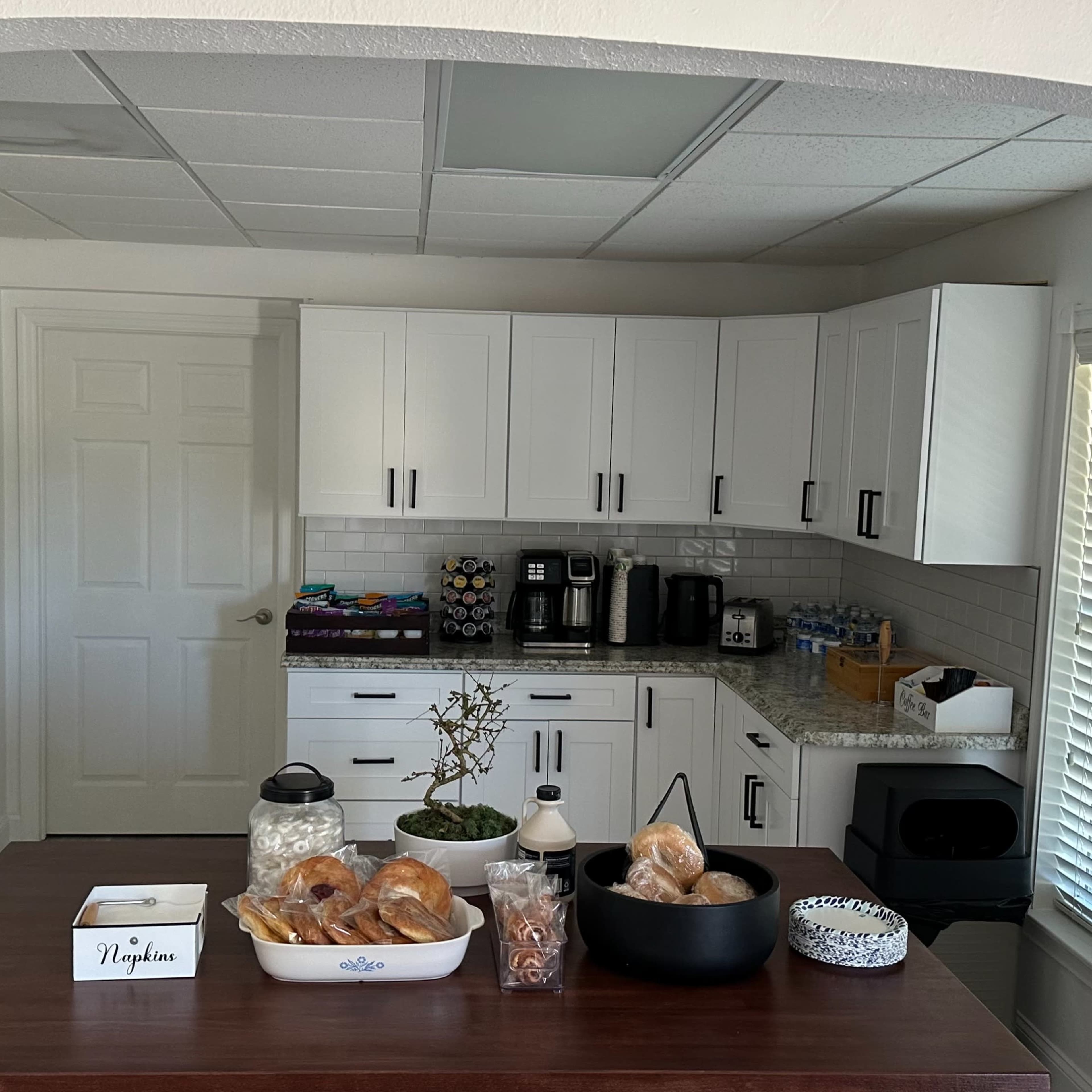 A modern kitchen with white cabinets, a countertop displaying baked goods, and various appliances arranged in a well-lit space.