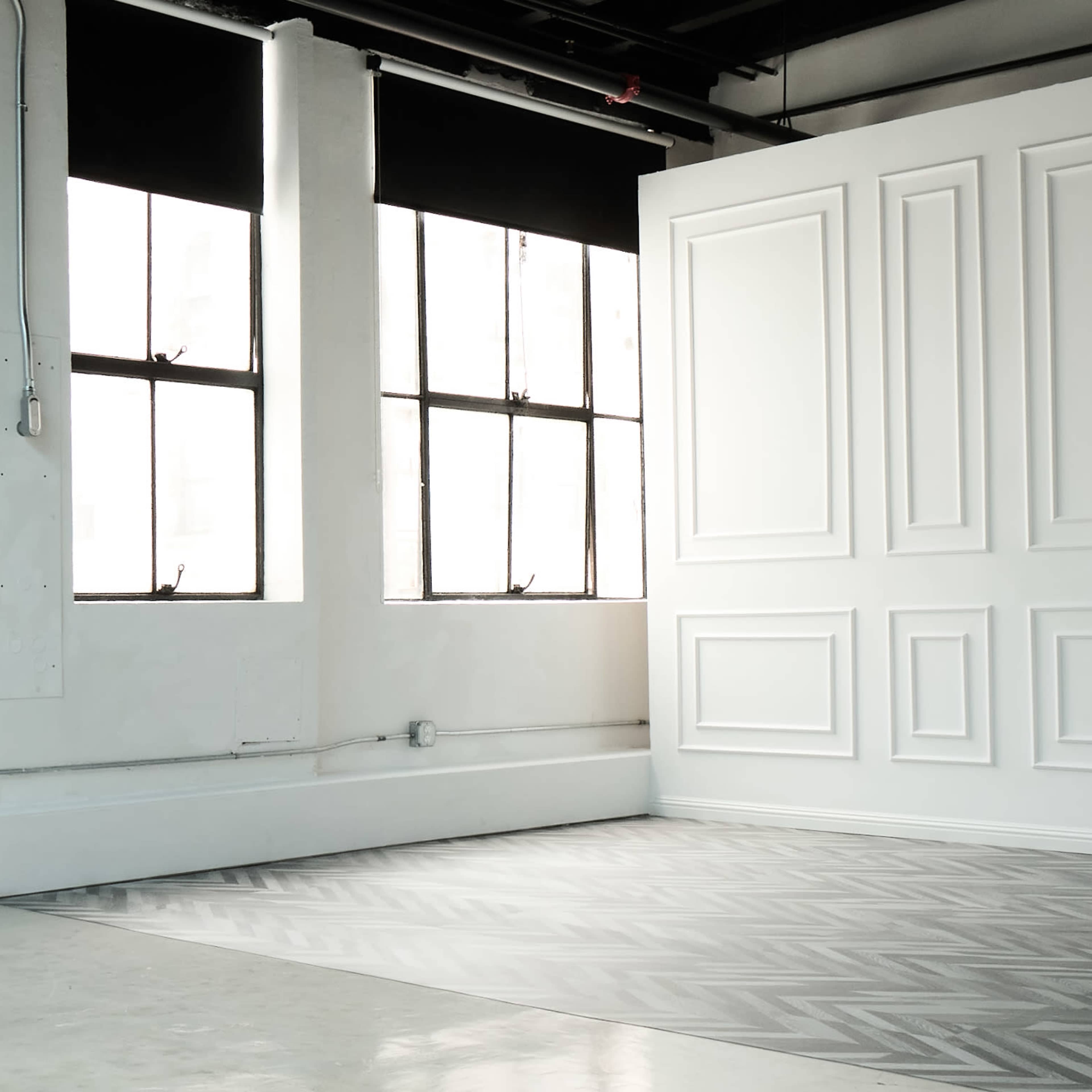 A brightly lit room with two large windows and a textured wall featuring rectangular paneling.