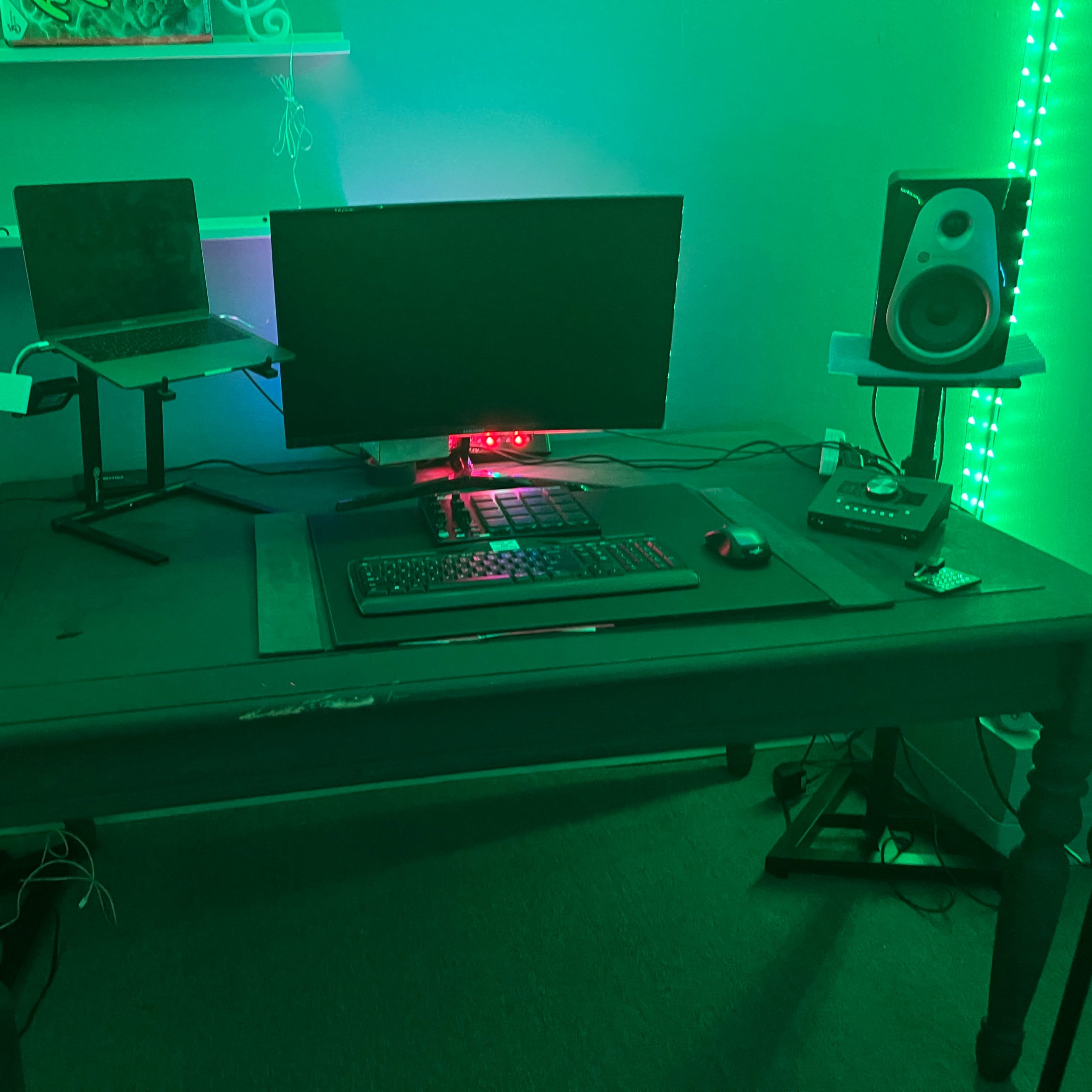 A desk setup features a computer monitor, a laptop on a stand, speakers on either side, and various electronic devices, all illuminated by green lighting.