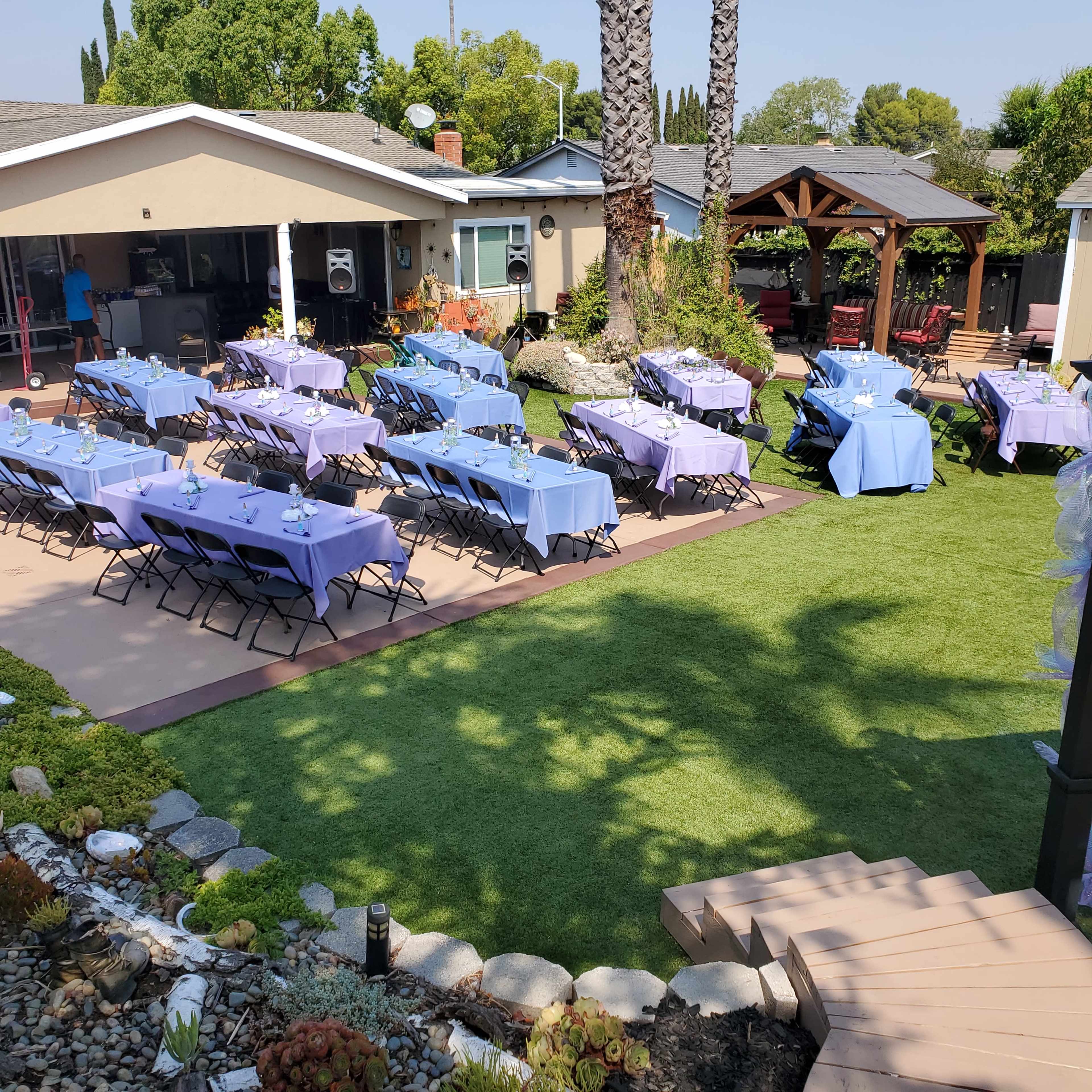 The scene shows a backyard setting with rows of tables covered in purple tablecloths, arranged on a patio surrounded by greenery and palm trees.