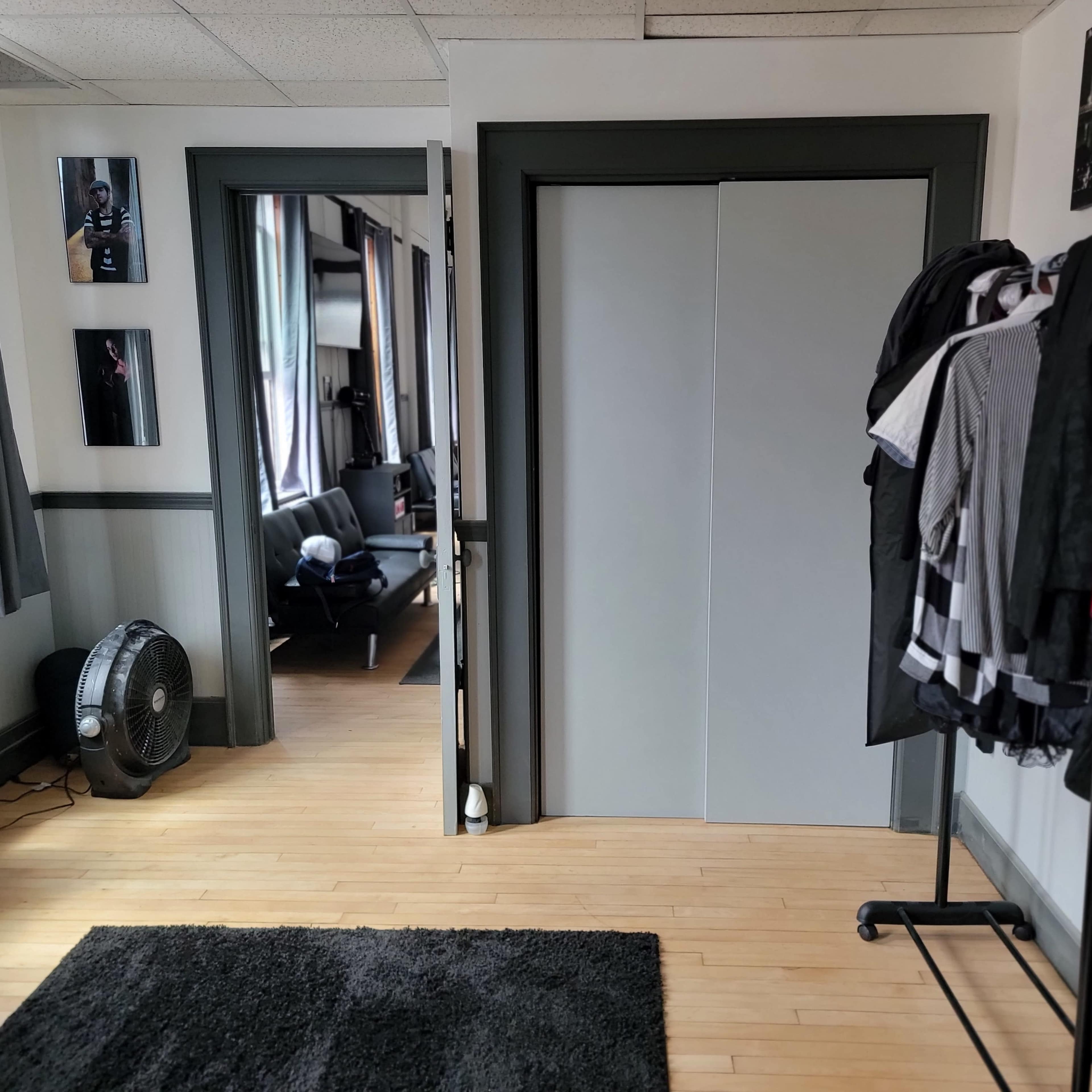 An interior room with a clothing rack holding several garments, a fan on the floor, and two doorways leading to other areas.