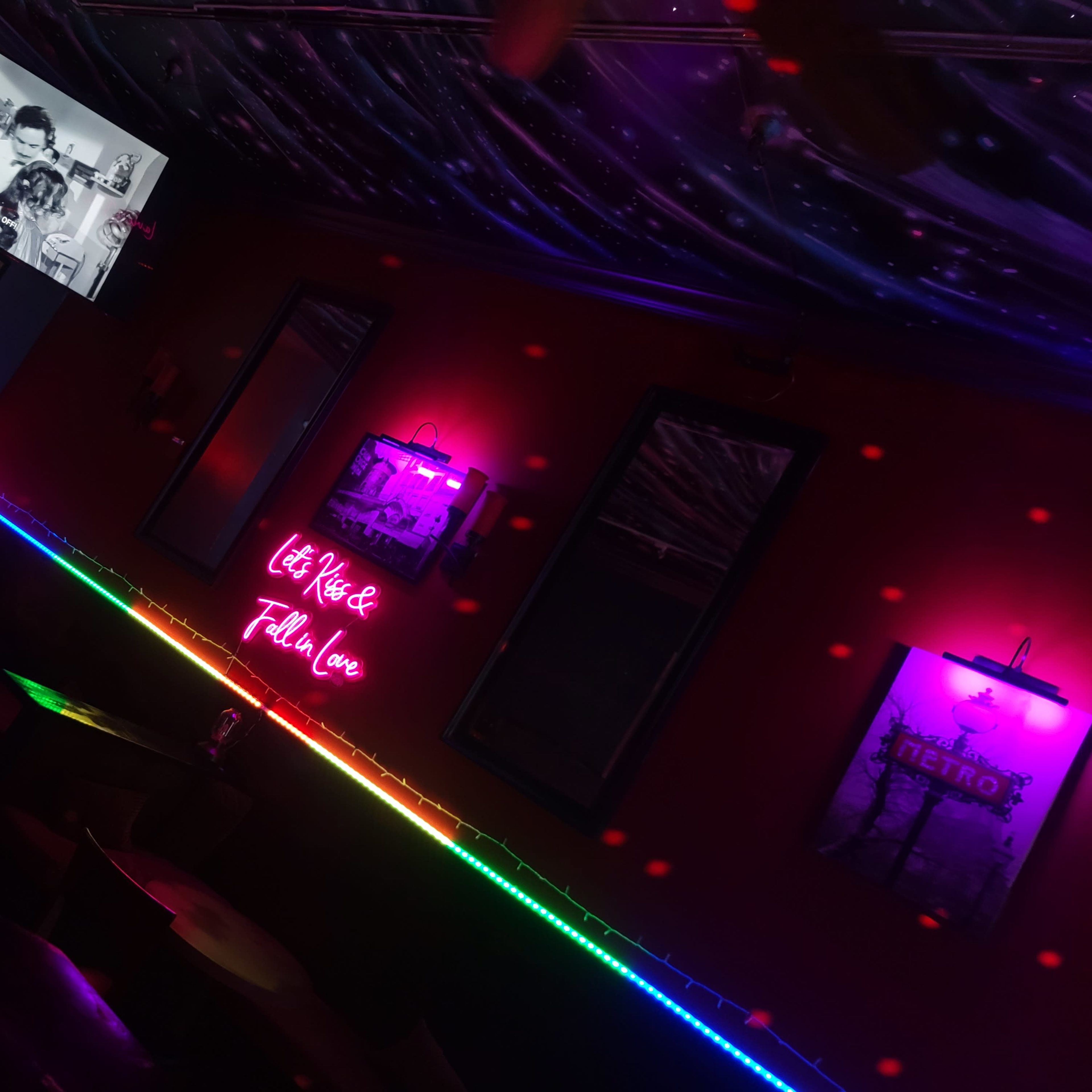 The interior of a dimly lit bar features a neon sign and a television displaying a black-and-white image, with colored lights lining the edges of the seating area.