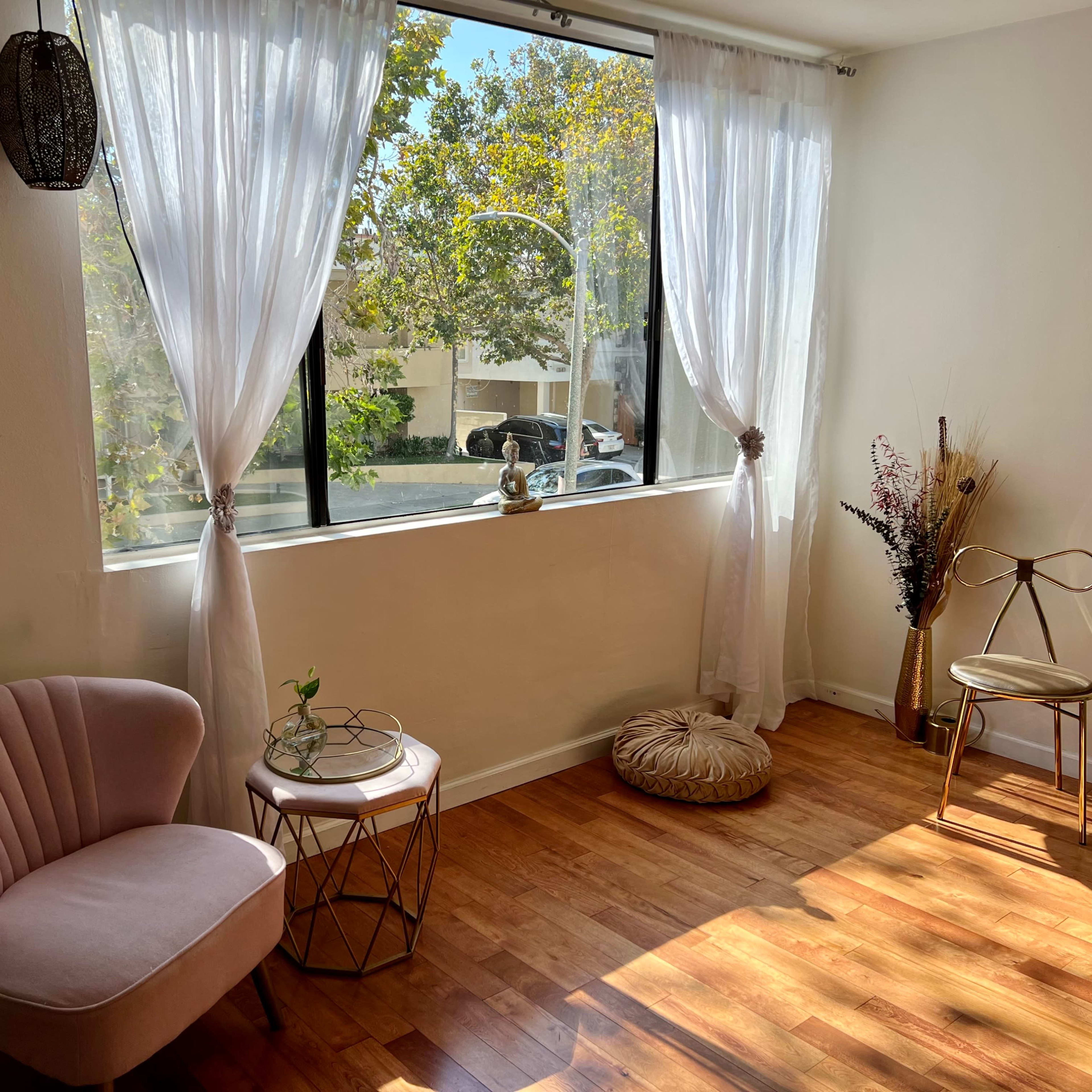 A well-lit room with large windows, a pair of chairs, a small table, and decorative plants.