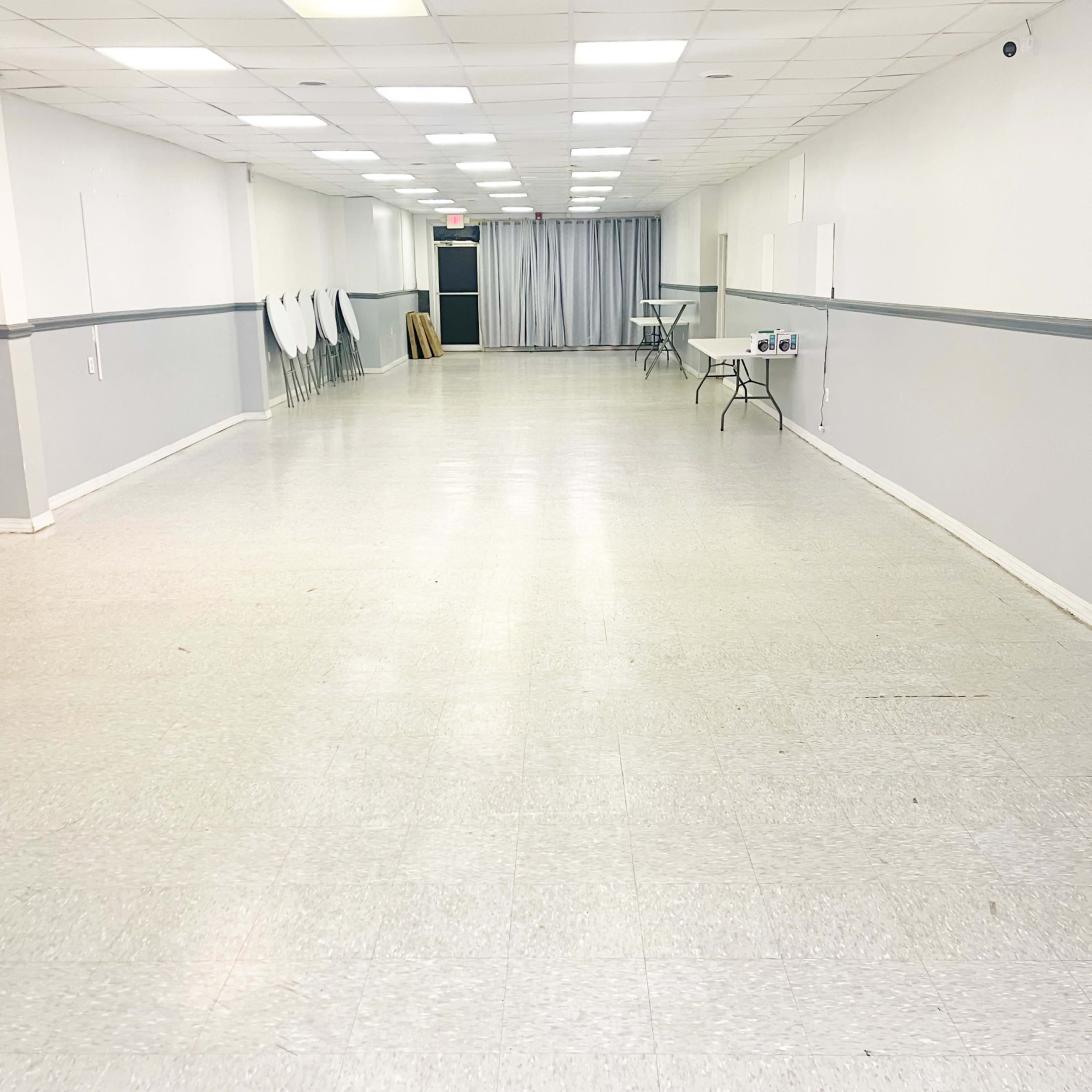 A large, empty hall with white walls, a gray stripe, and rows of stacked chairs along one side.