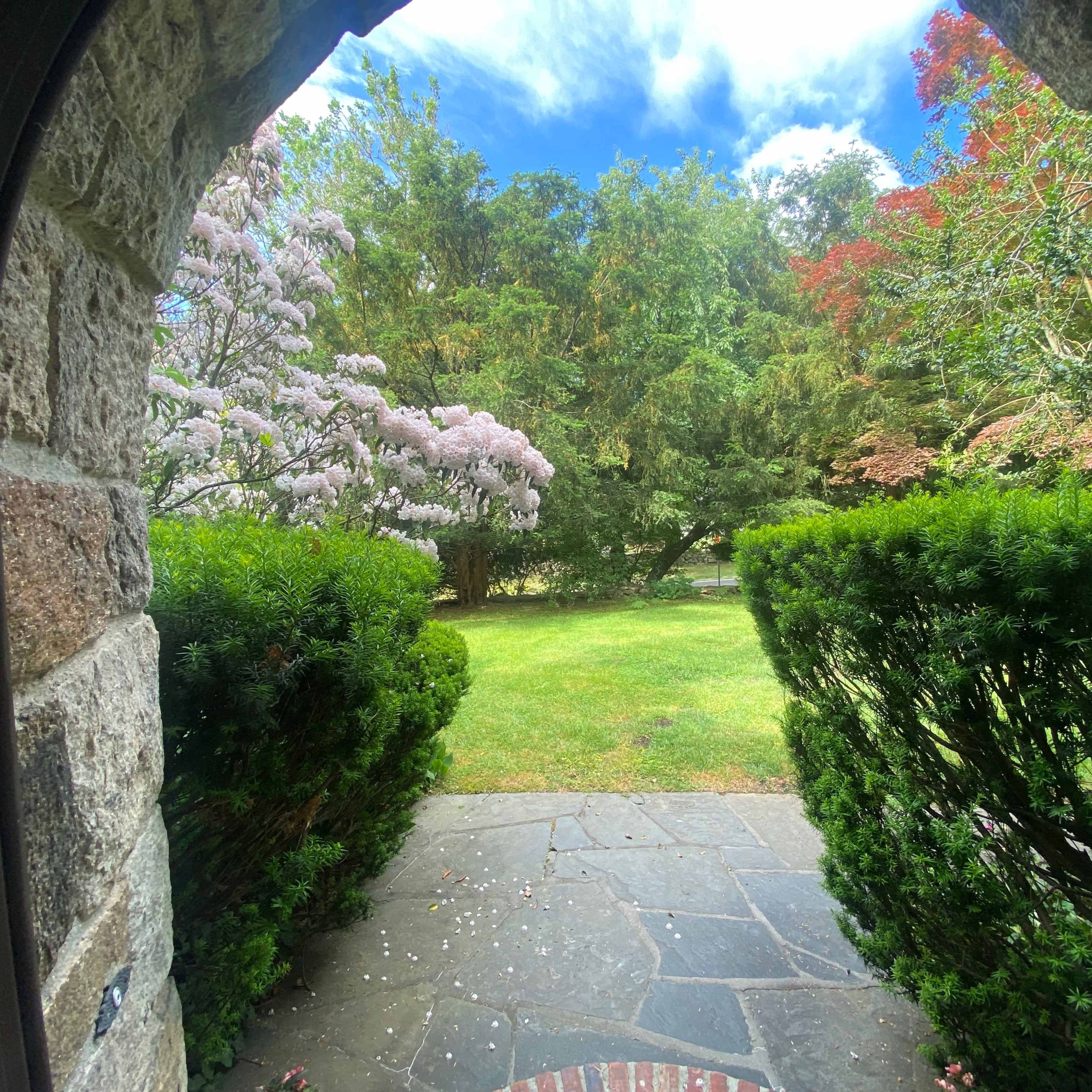 The view from the stone archway reveals a green garden with flowering trees and shrubs under a partly cloudy sky.