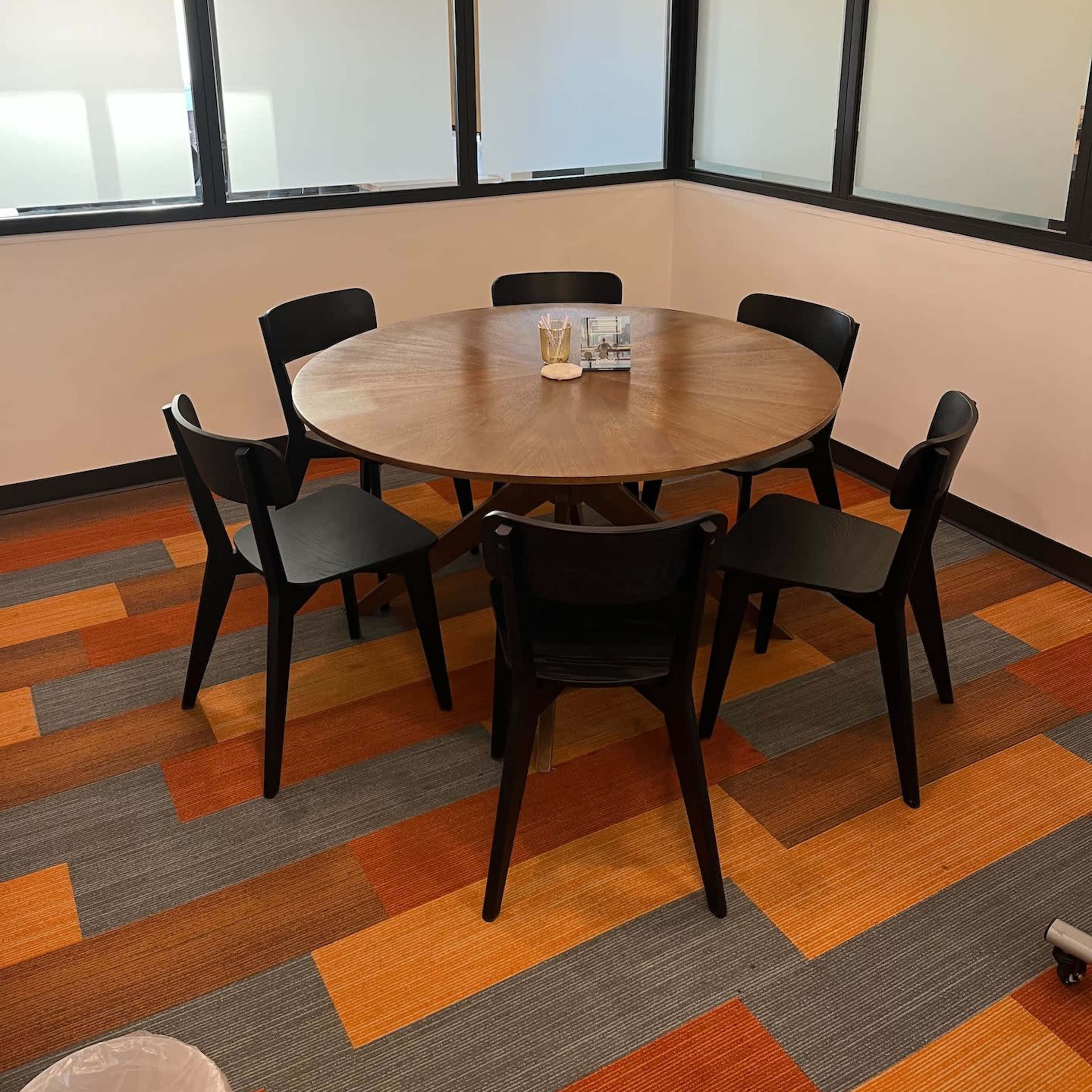 A round wooden table surrounded by six black chairs is positioned in a corner office with large windows.