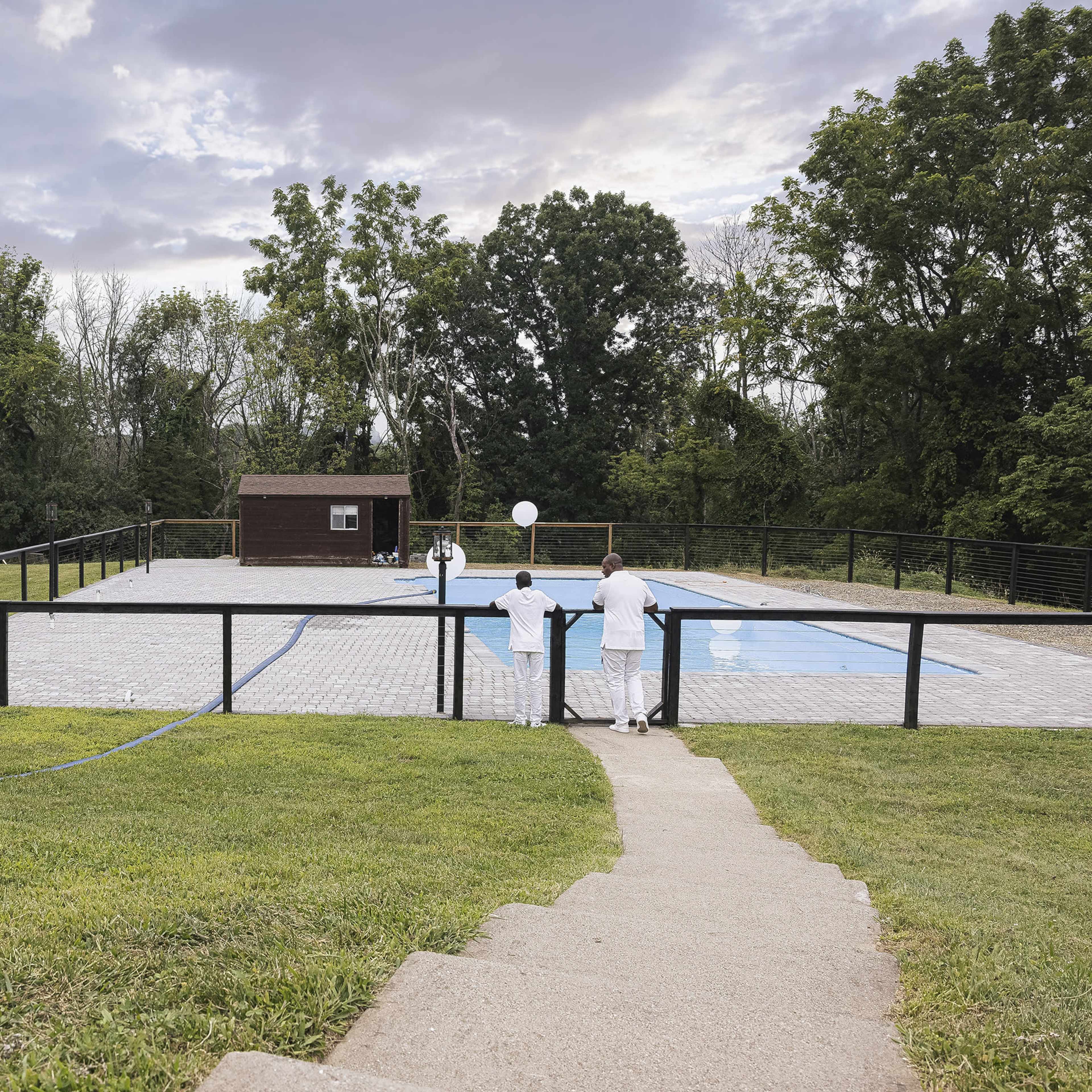 A paved area with an empty swimming pool is surrounded by grass and trees, with two people standing near the edge of the pool.