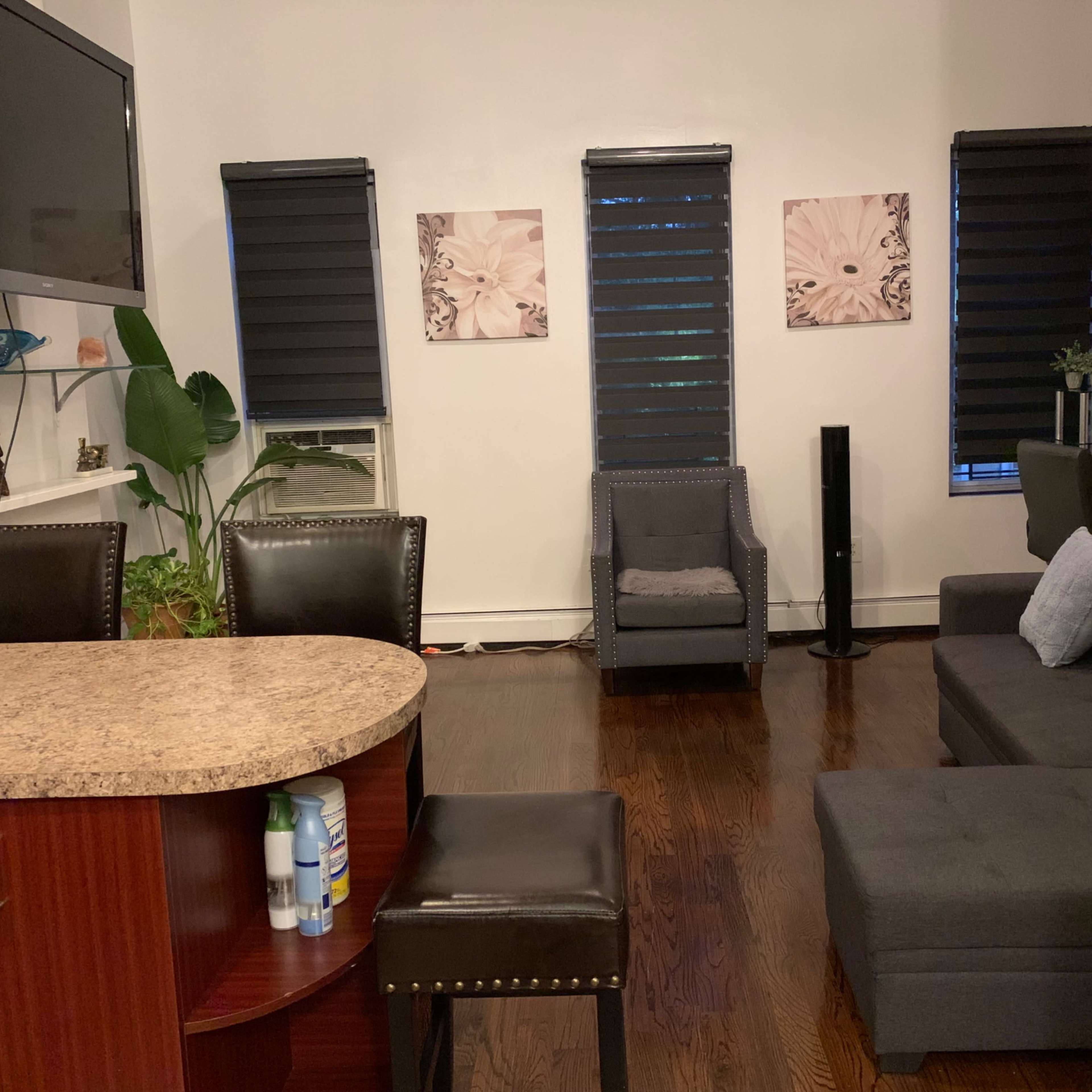 A modern living space with a kitchen bar, two black blinds-covered windows, a gray sectional sofa, and two decorative wall prints.