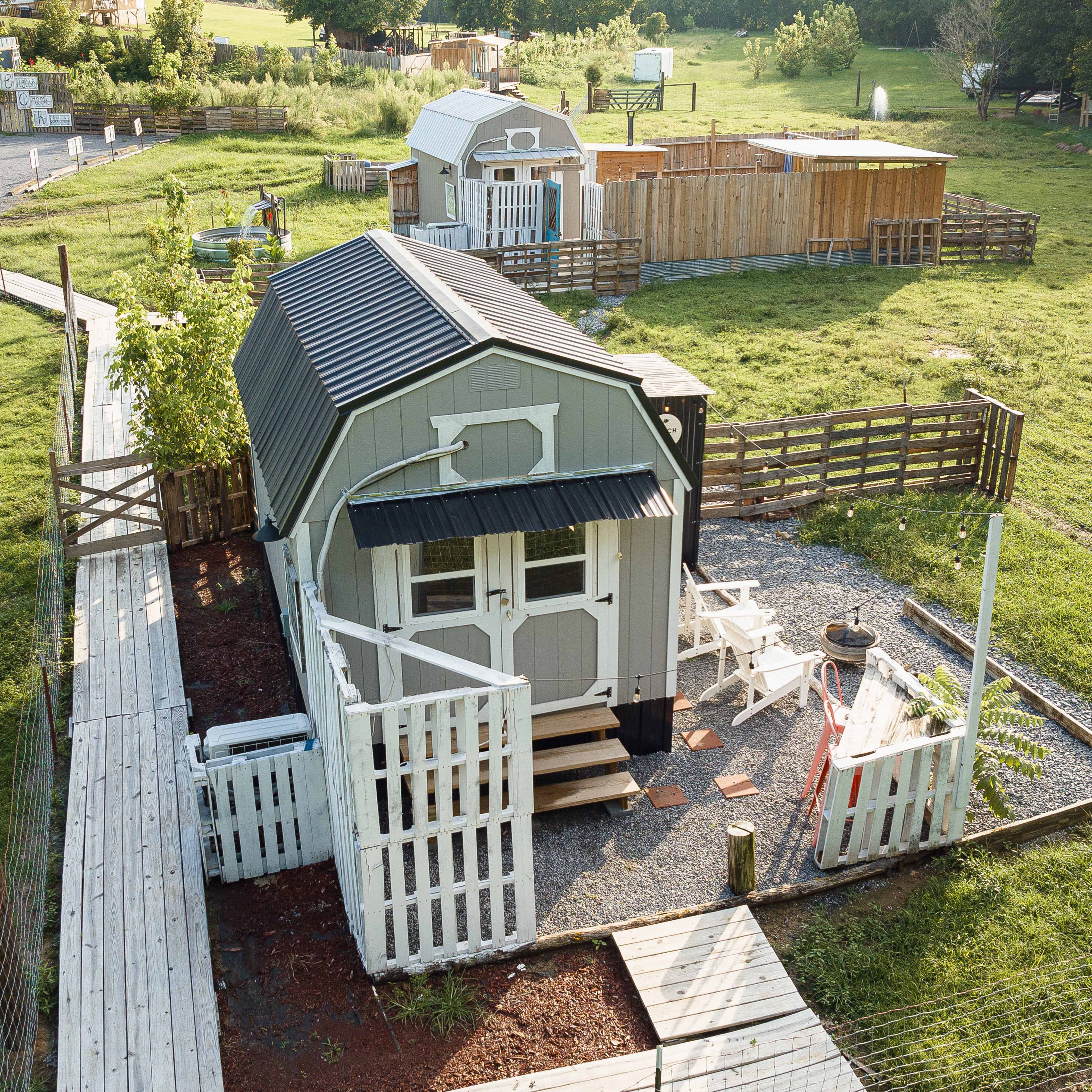 A small, modern cabin surrounded by a wooden fence and a gravel pathway, with additional structures and greenery in the background.