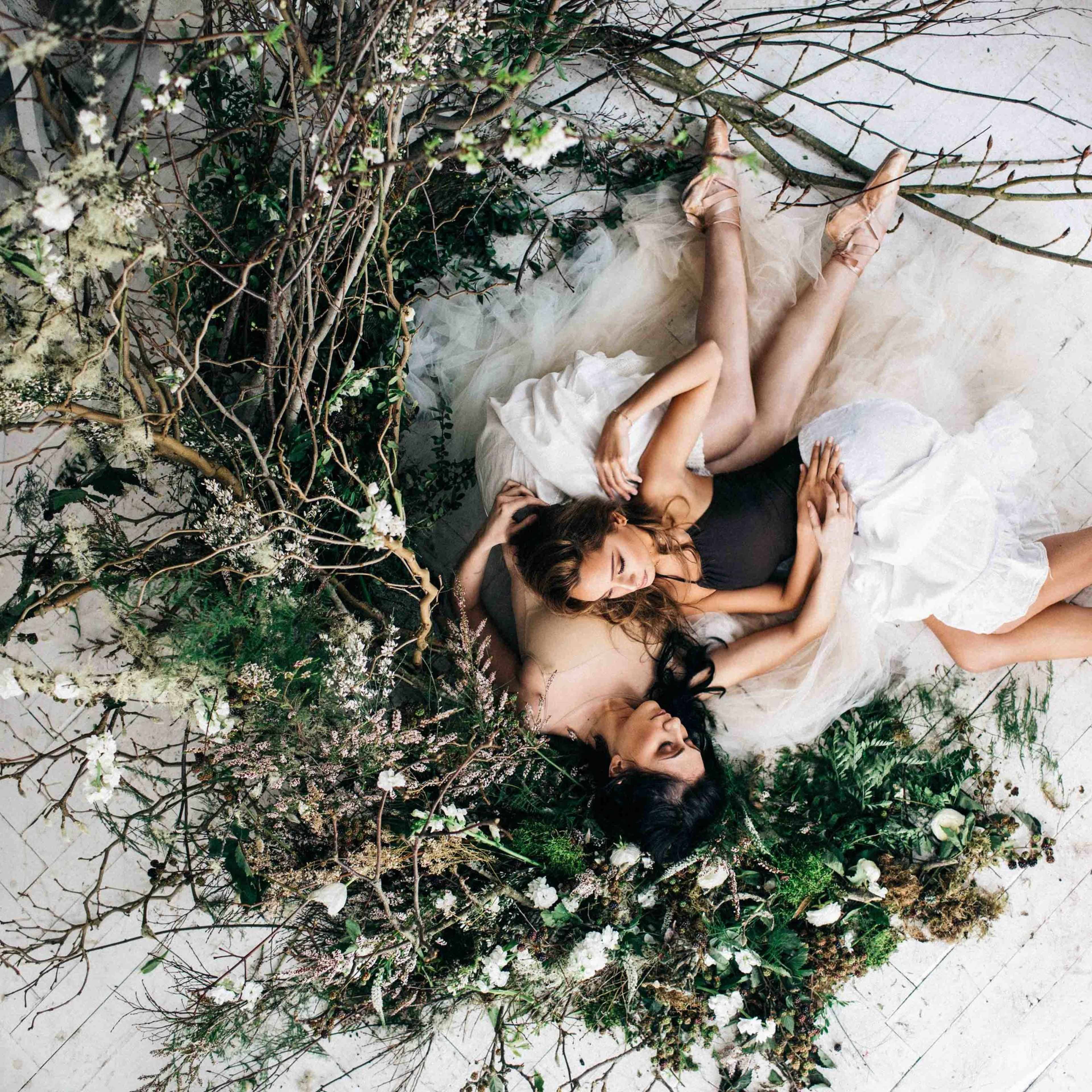 Two dancers in tutus rest on a bed of greenery and branches, surrounded by white flowers and soft fabric on a light floor.