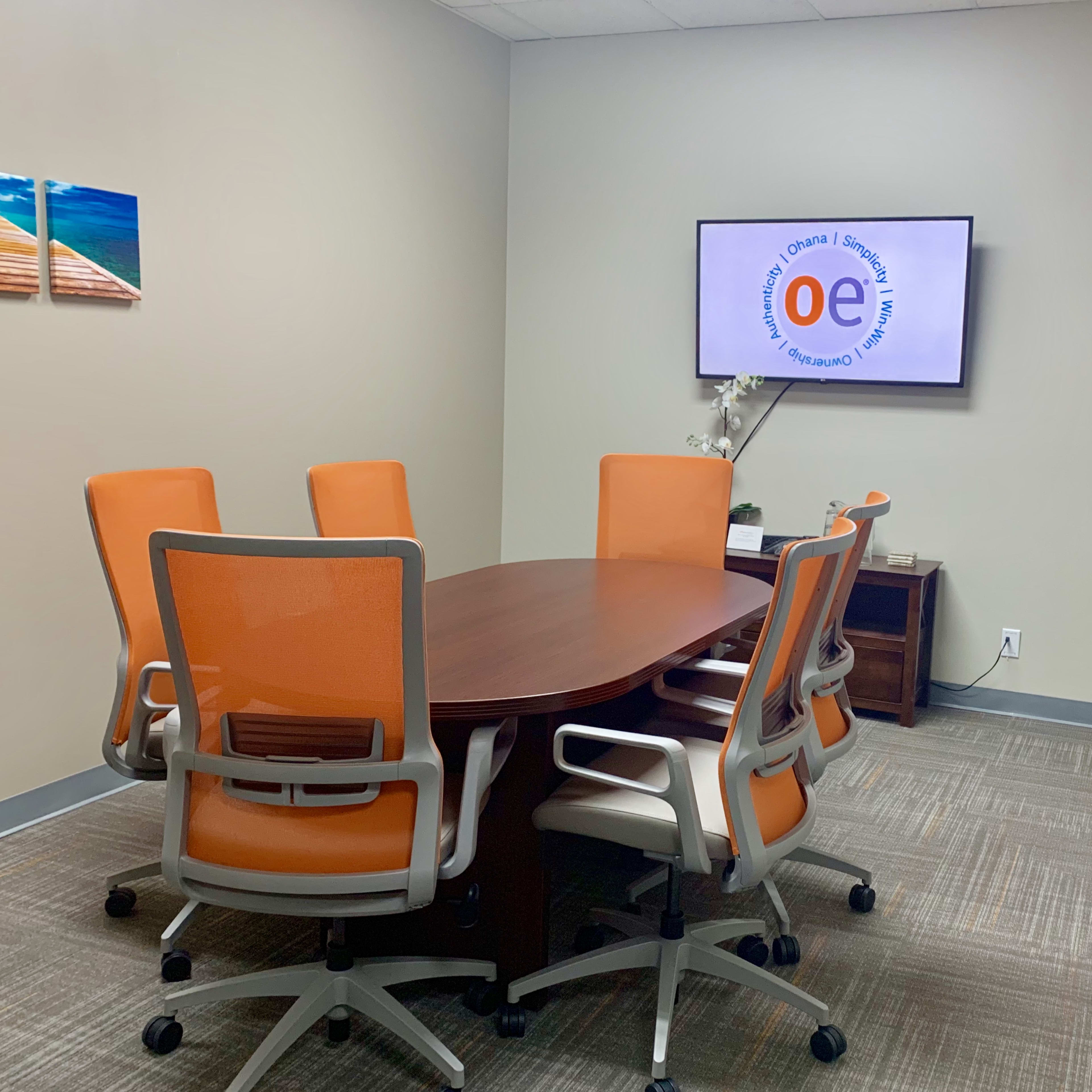 A conference room is depicted with a round wooden table surrounded by six orange chairs, a television displaying a logo, and a whiteboard on the wall.
