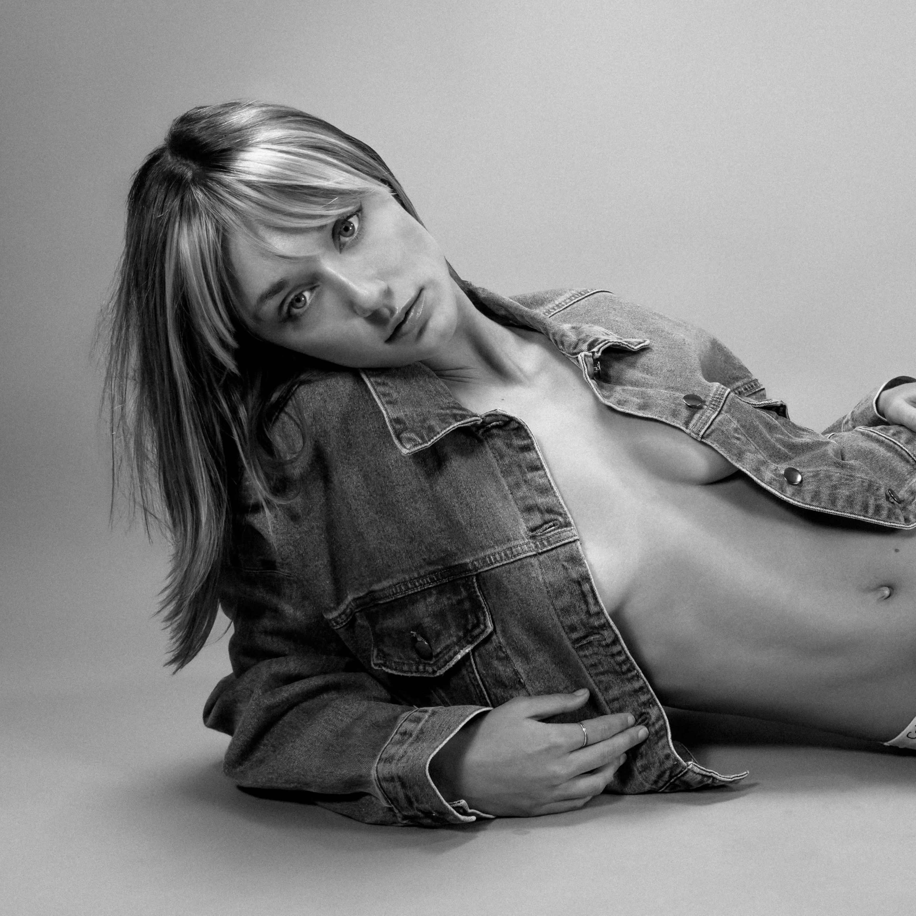 A person lies on a plain background, wearing a denim jacket and underwear, with one arm resting on their side.