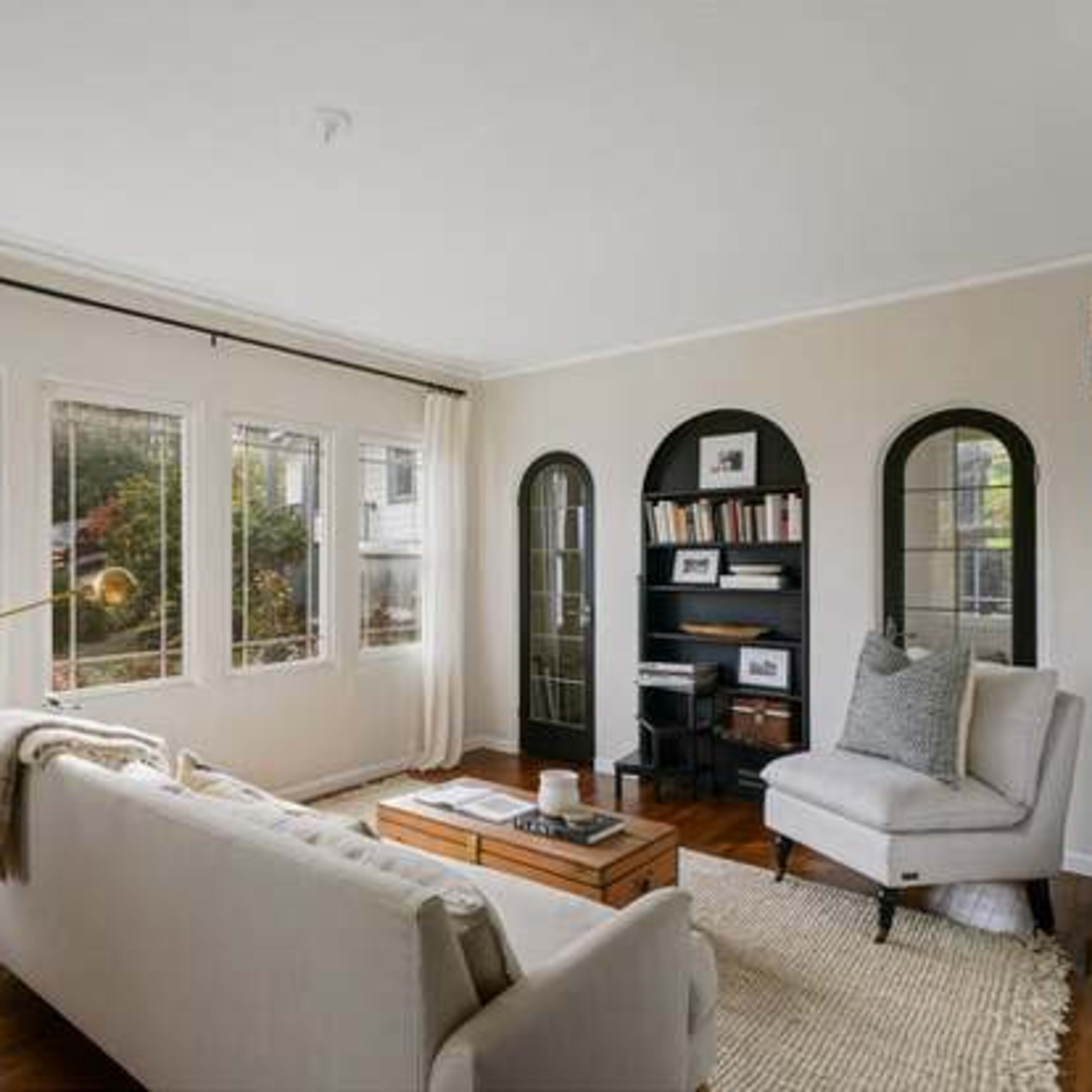 A cozy living room features a white sofa, a wooden coffee table, and large windows allowing natural light, with bookshelves and decorative arches in the background.
