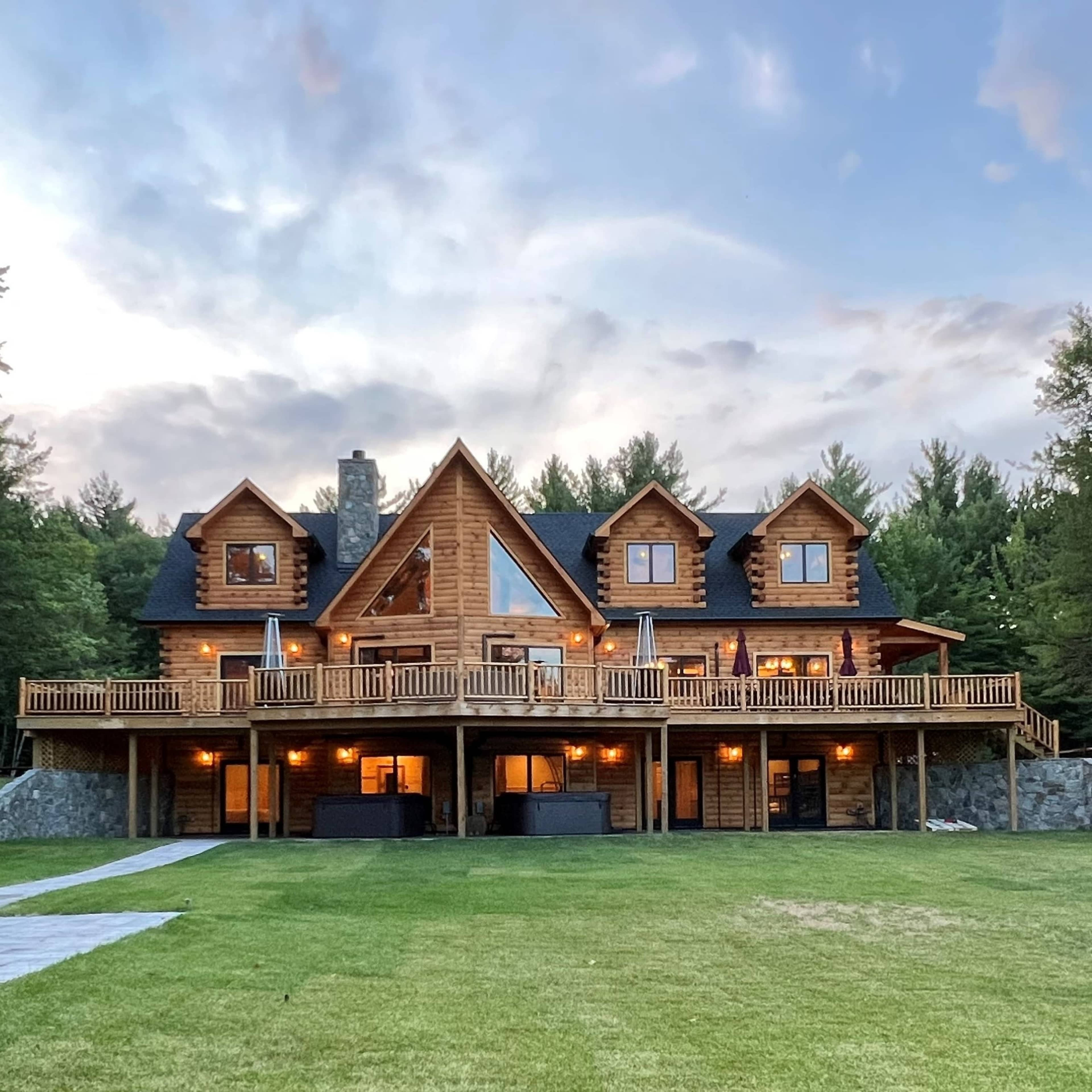 A large wooden house with multiple gabled roofs is situated on a grassy lawn surrounded by trees, featuring a spacious deck and stone landscaping.