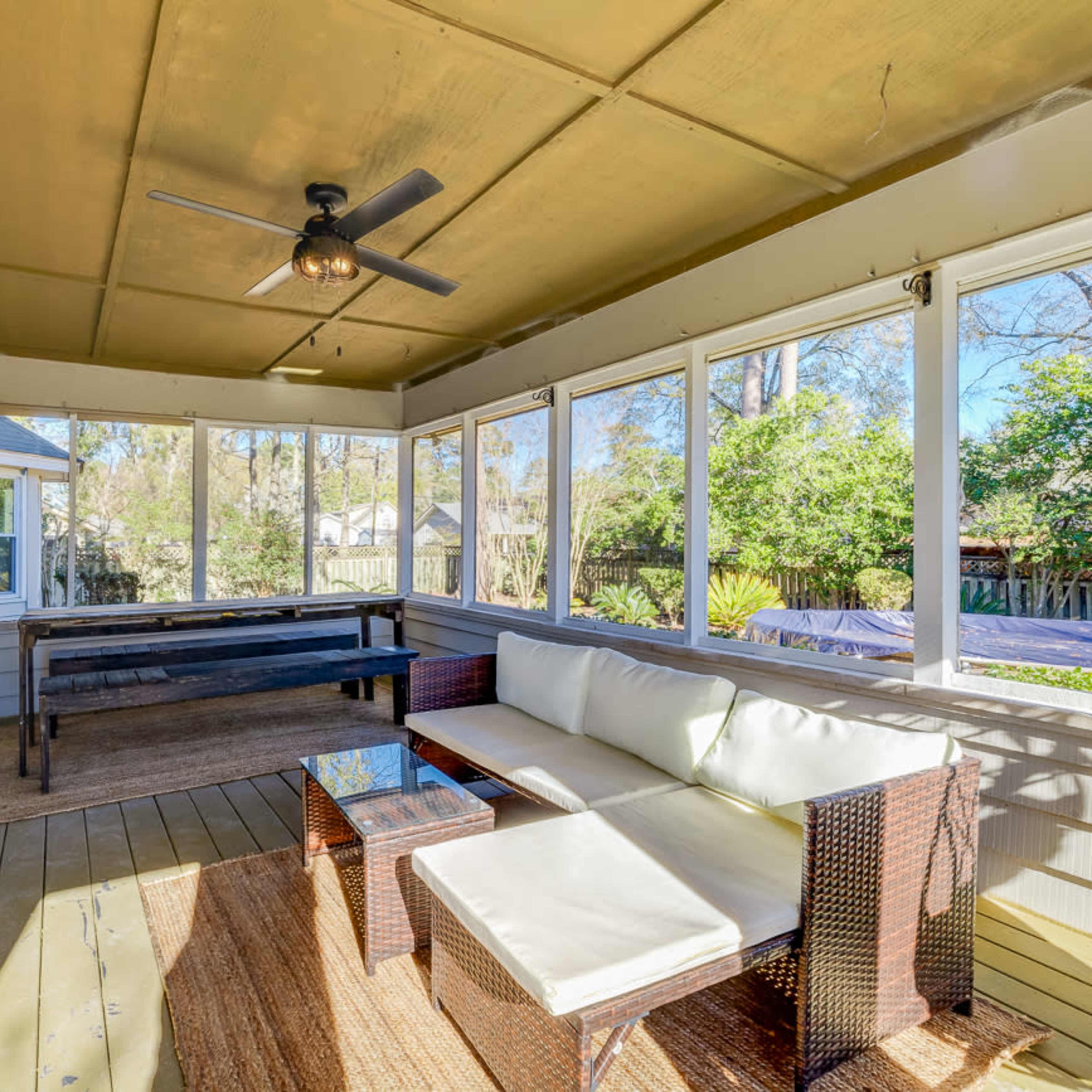 A screened-in porch with a seating area, a coffee table, and a ceiling fan, surrounded by greenery.