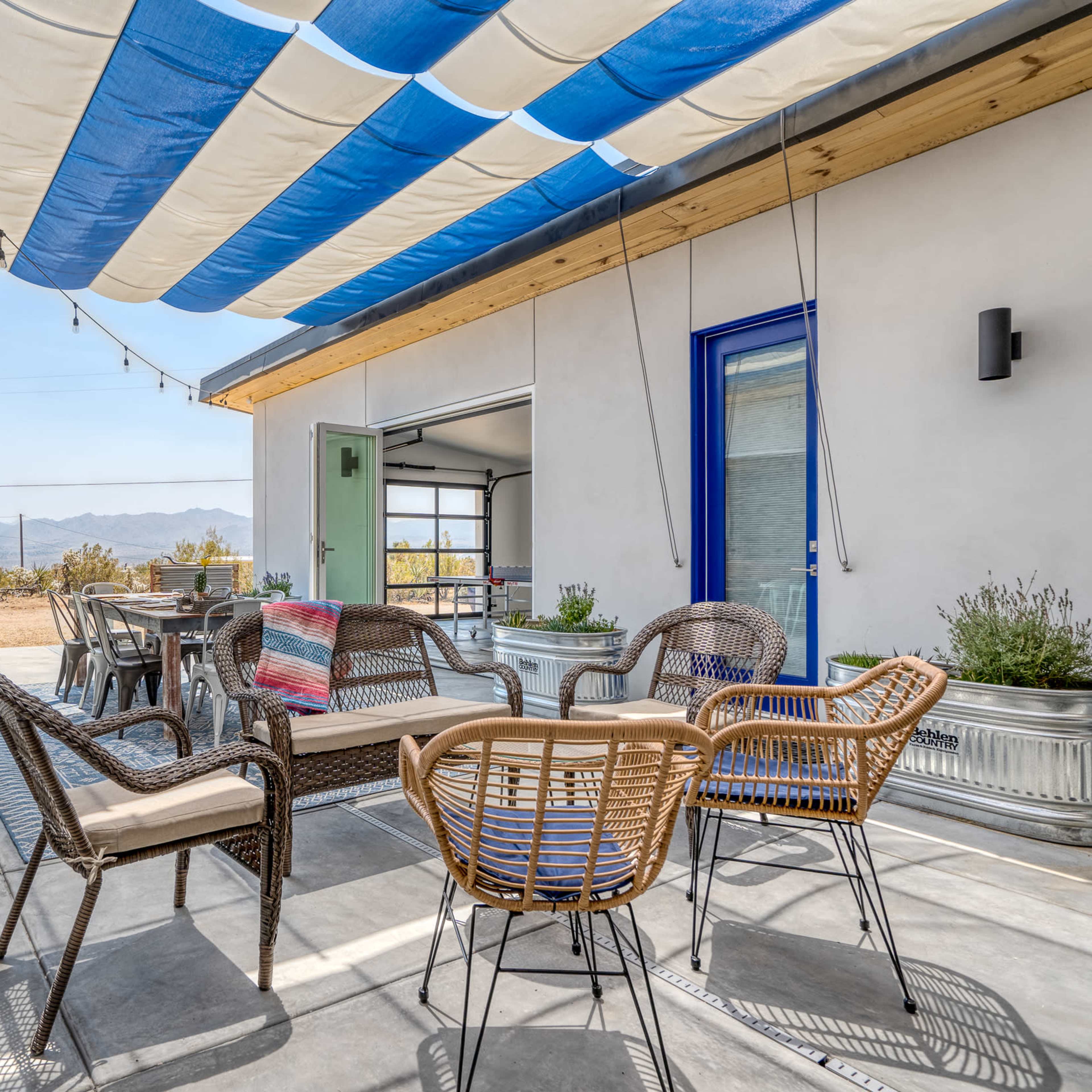 A patio area with a striped awning, featuring several chairs and a small table, surrounded by potted plants and a view of mountains in the distance.