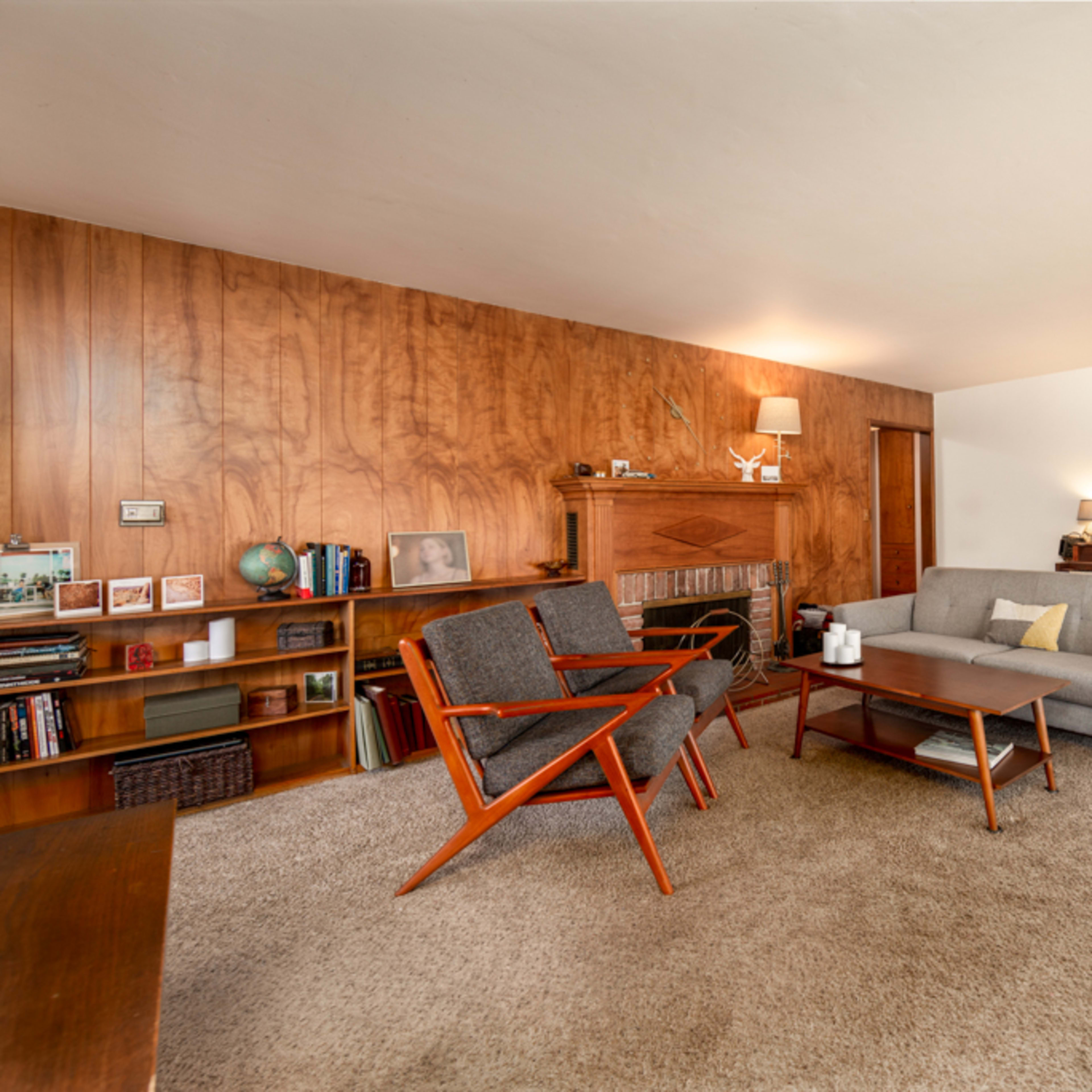 A mid-century modern living room with wooden paneling, a gray sofa, vintage chairs, and a styled bookshelf against one wall.