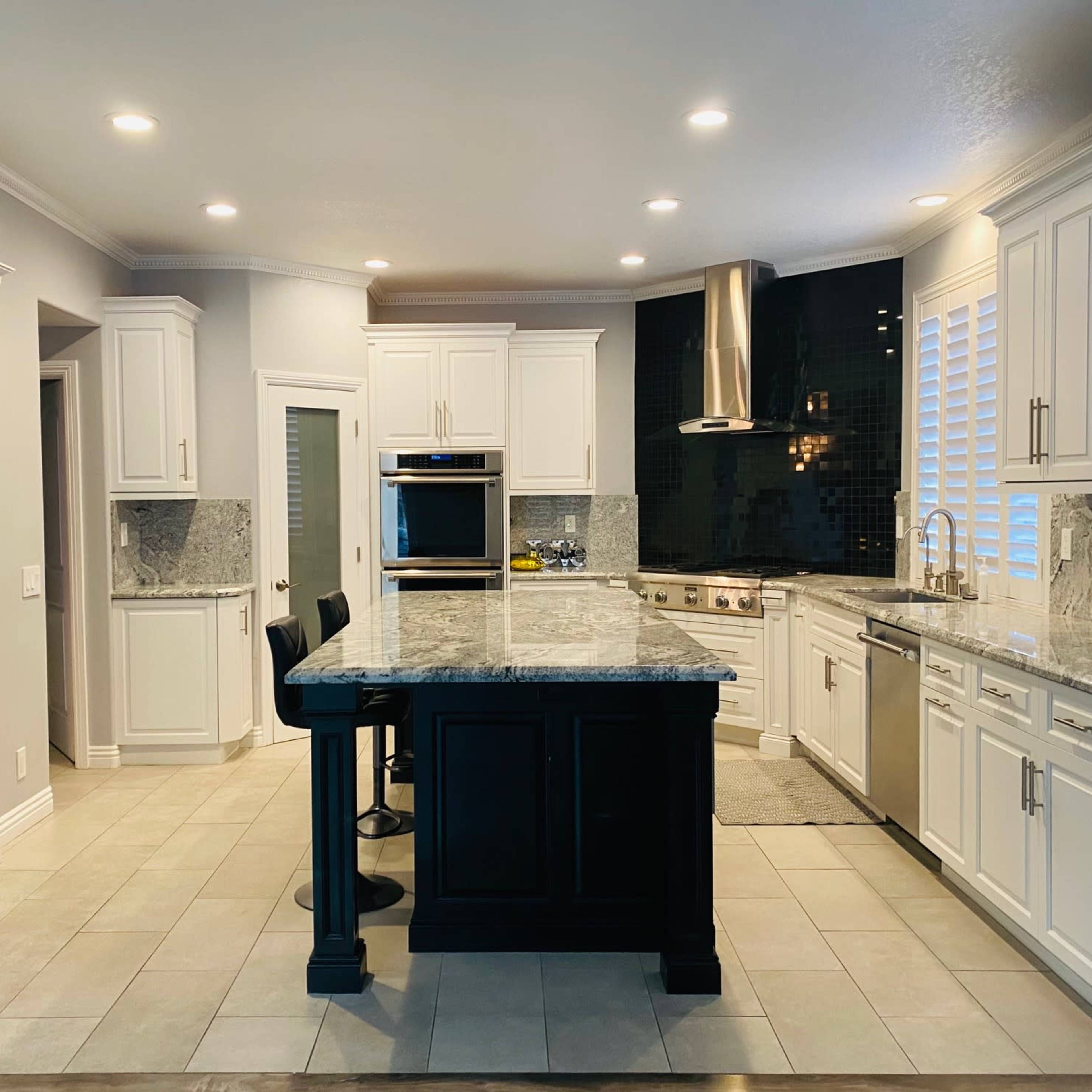 A modern kitchen features white cabinetry, a dark island with seating, stainless steel appliances, and a marble countertop.