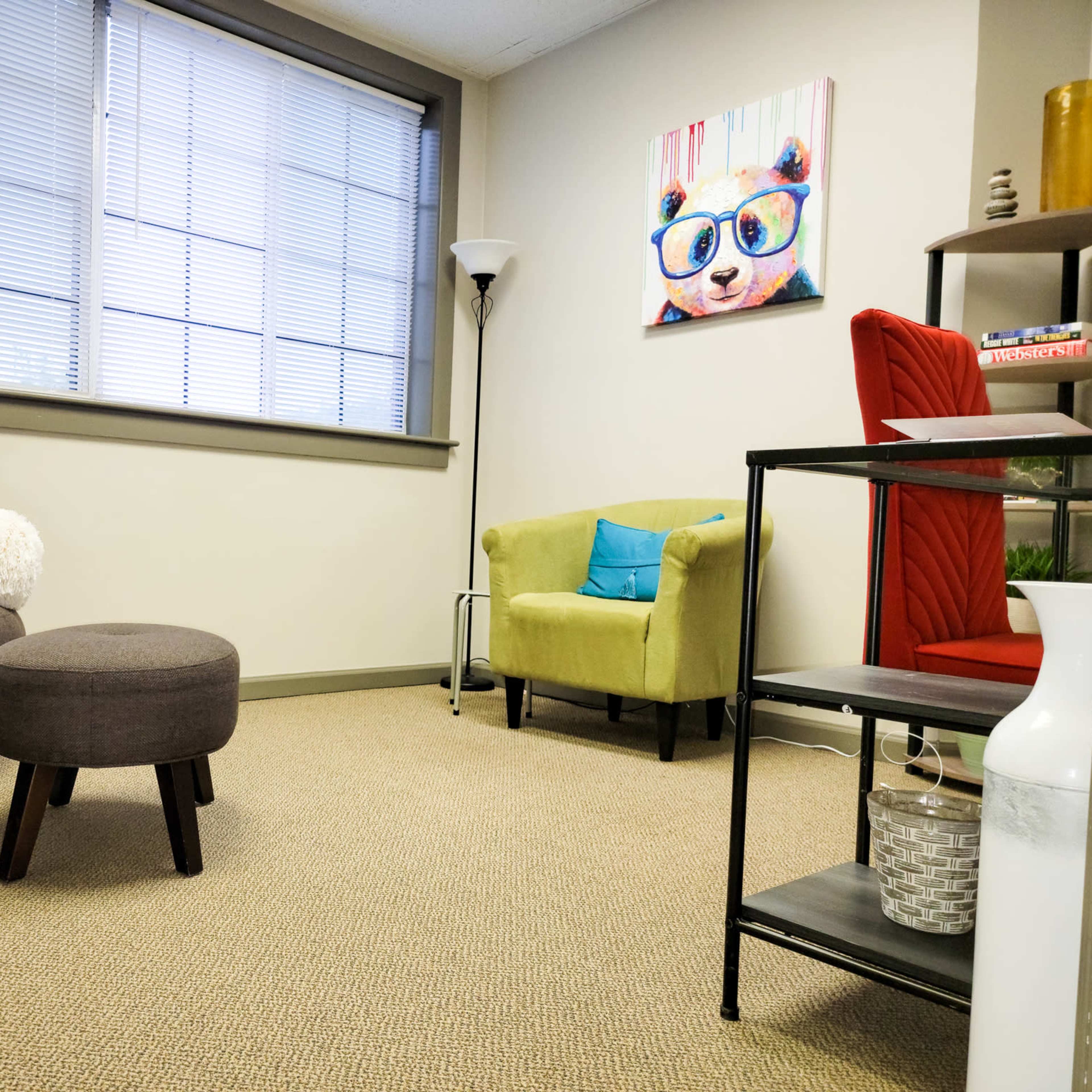 A cozy room with two armchairs, a couch, and a small round ottoman, all arranged on a carpeted floor beneath bright windows.