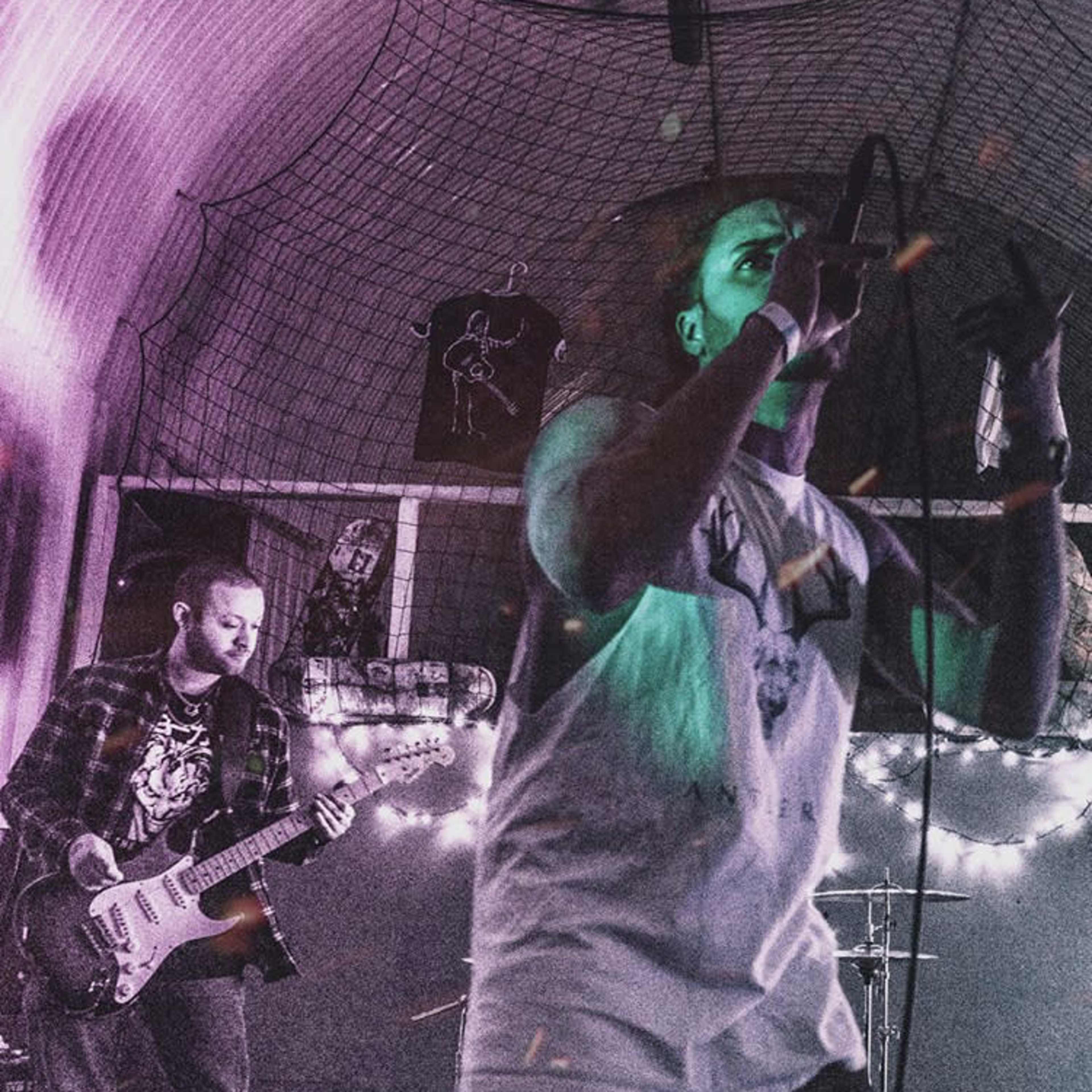 A vocalist performs on stage while a guitarist plays beside him in a dimly lit venue decorated with lights and band posters.