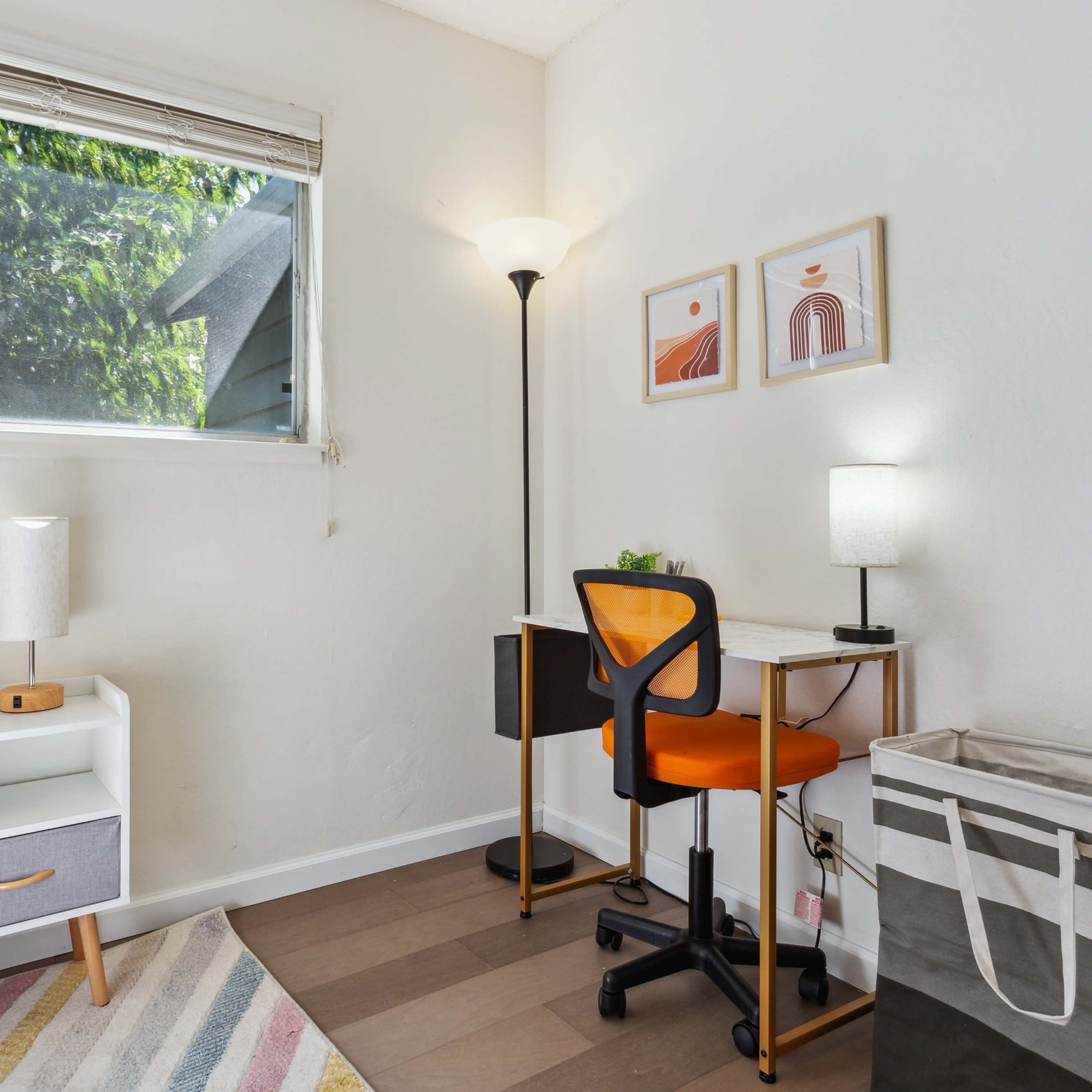 A small bedroom featuring a bed, a desk with an orange chair, and a window with natural light coming in, complemented by decorative framed art on the wall.