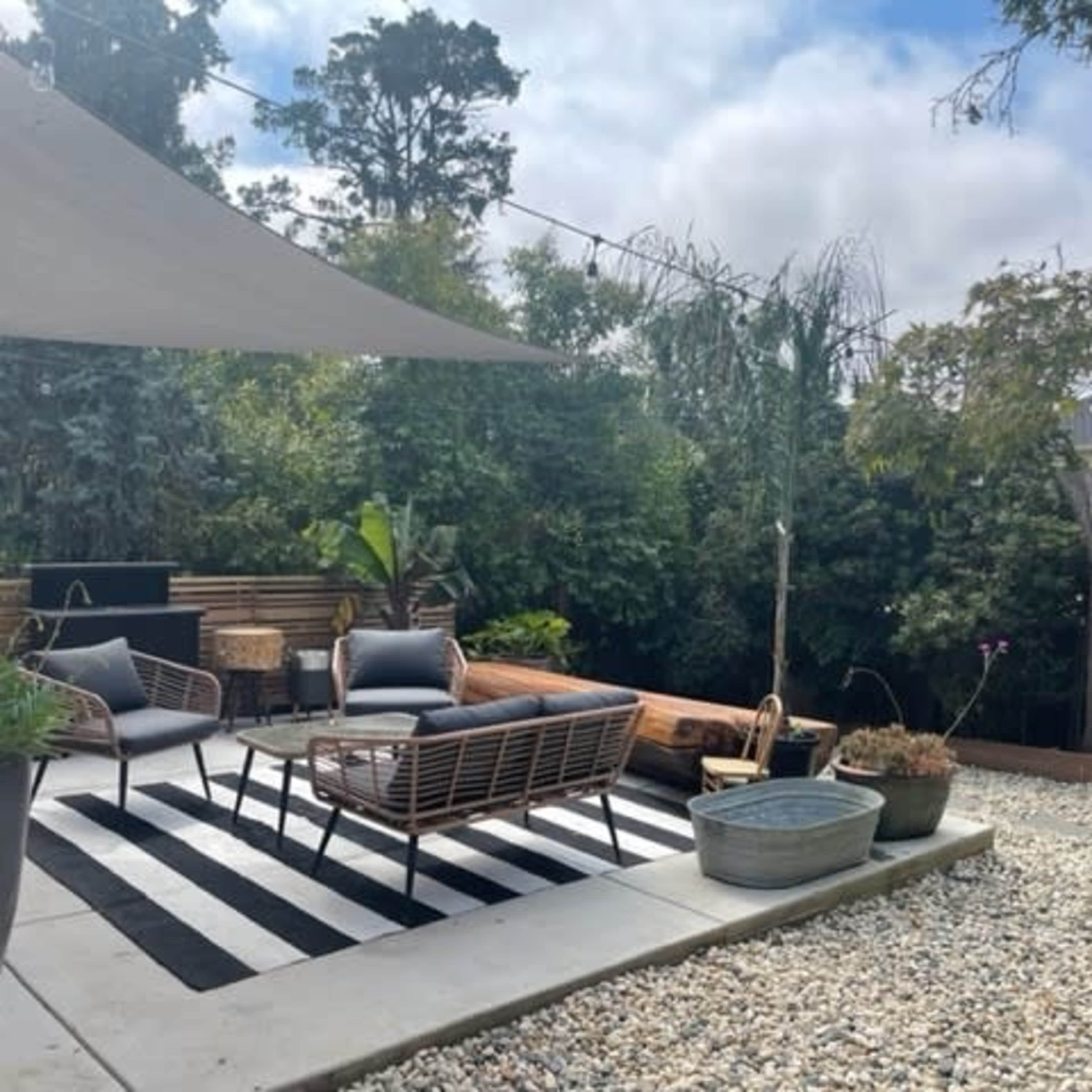 A patio area features outdoor seating arranged on a striped rug, surrounded by greenery and decorative planters.