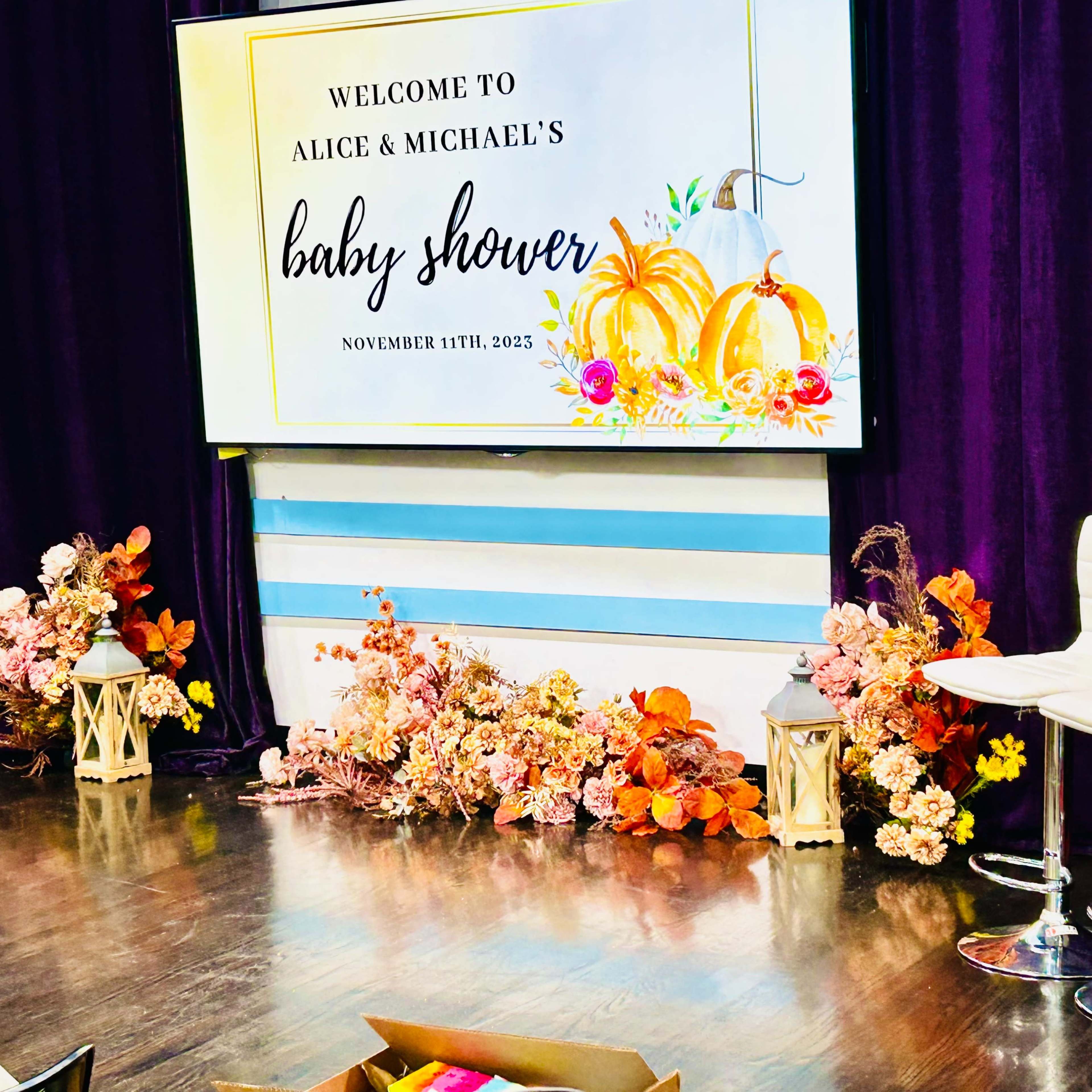 A decorated area for a baby shower, featuring a large display board with "Welcome to Alice & Michael's Baby Shower" and colorful floral arrangements at the base.