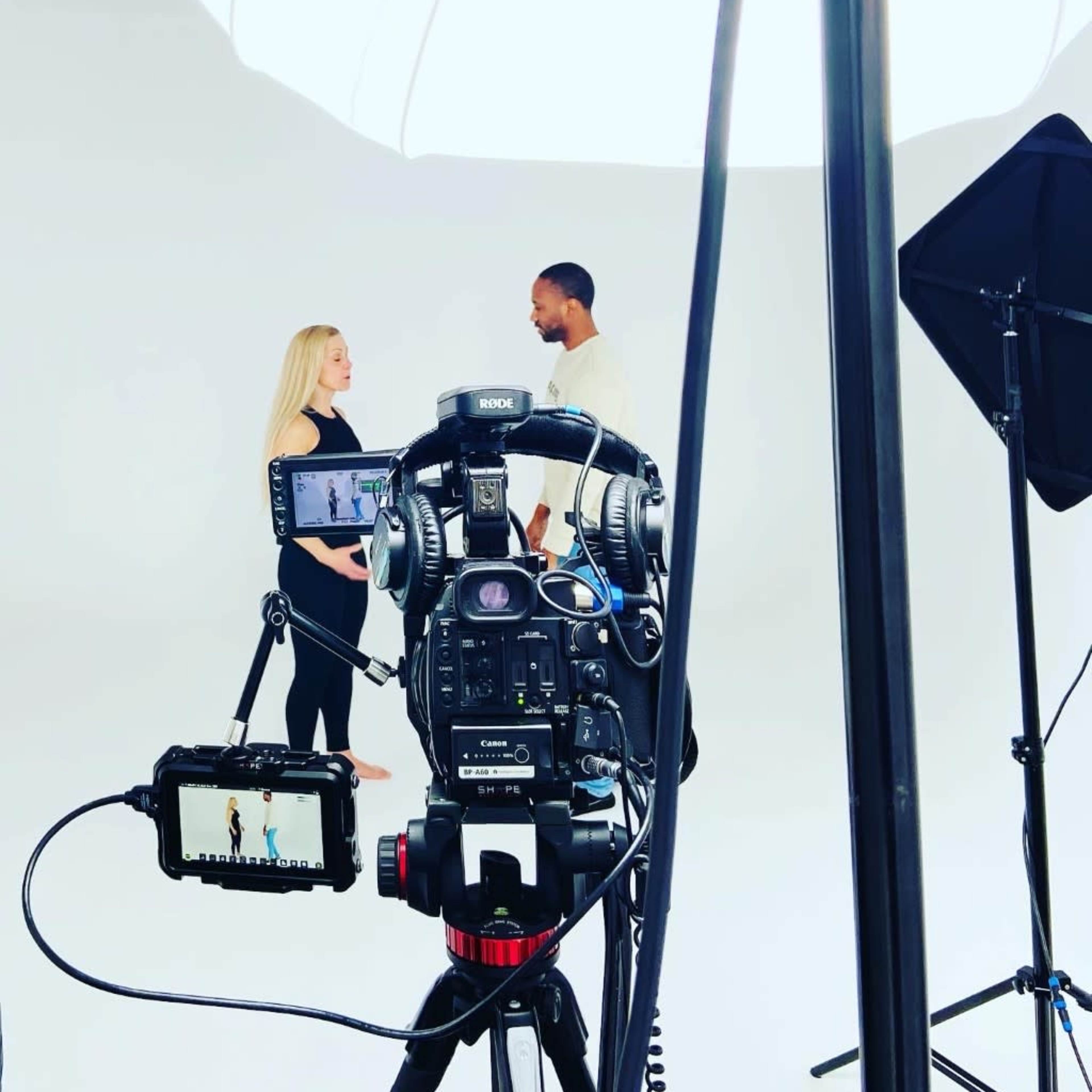 A camera set up on a tripod captures two individuals engaged in conversation in a well-lit studio against a plain white backdrop.