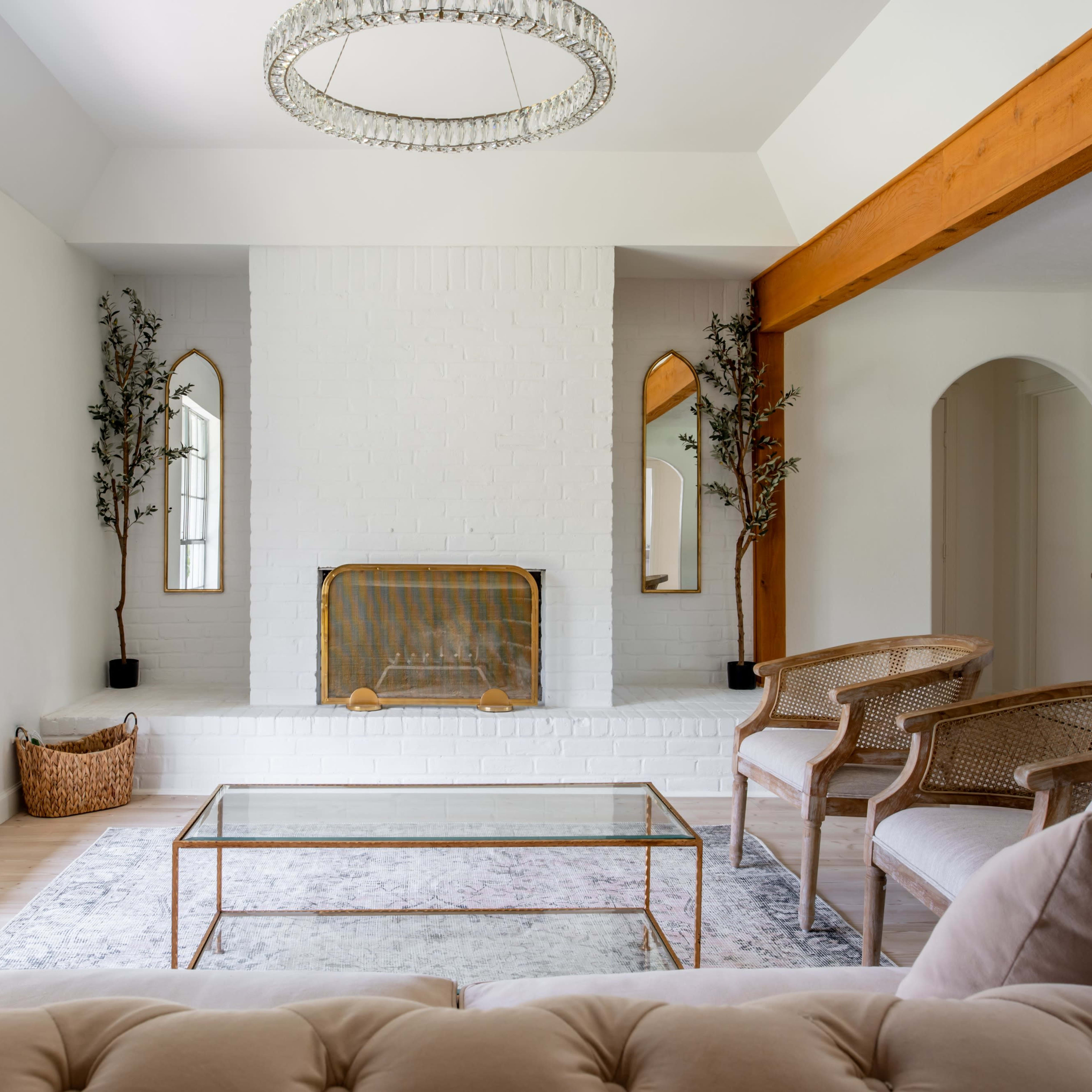 A bright living room features a white brick fireplace, a glass coffee table, and two rattan chairs, framed by large windows and decorative plants.