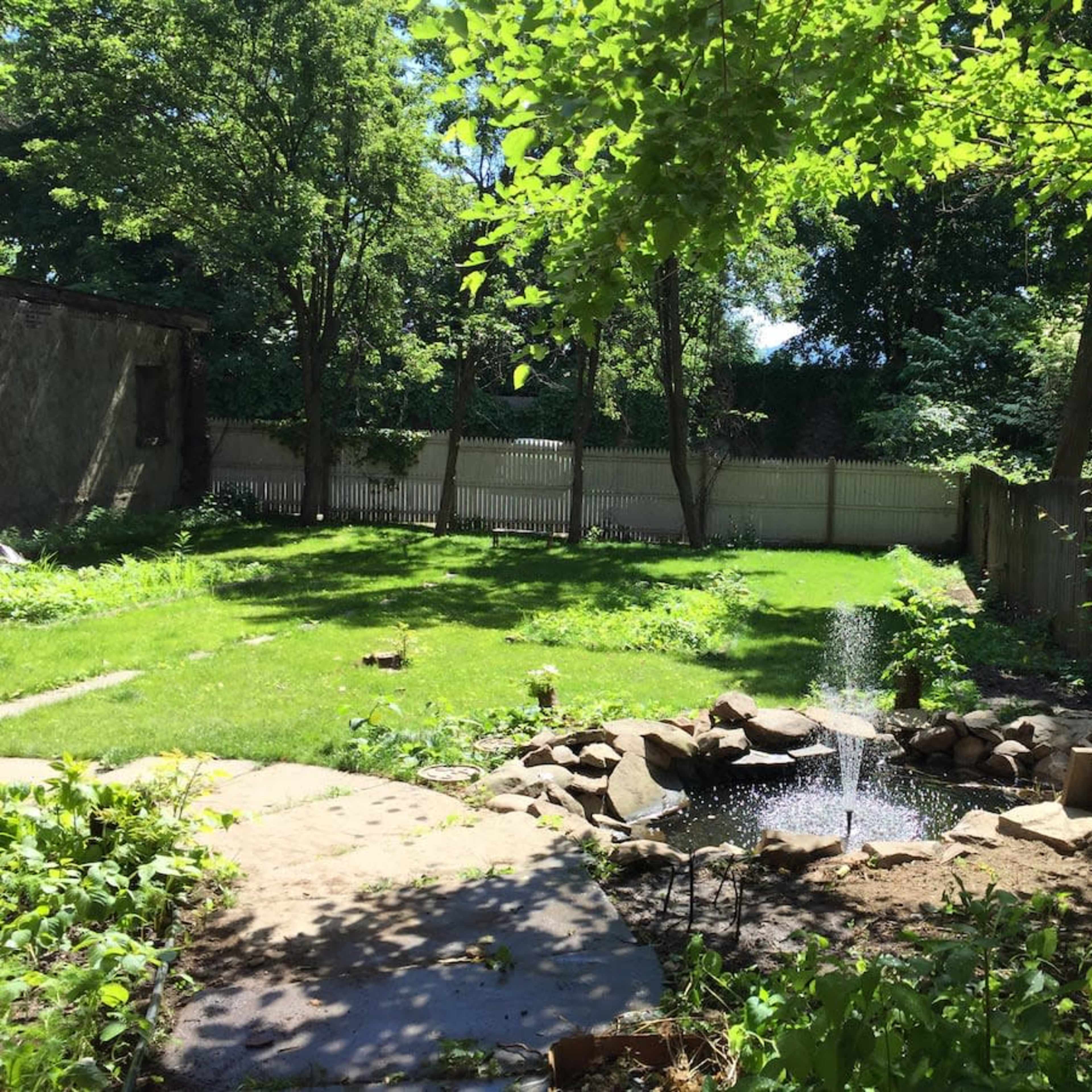 A small garden features a fountain surrounded by grass and rocks, with trees and a wooden fence in the background.