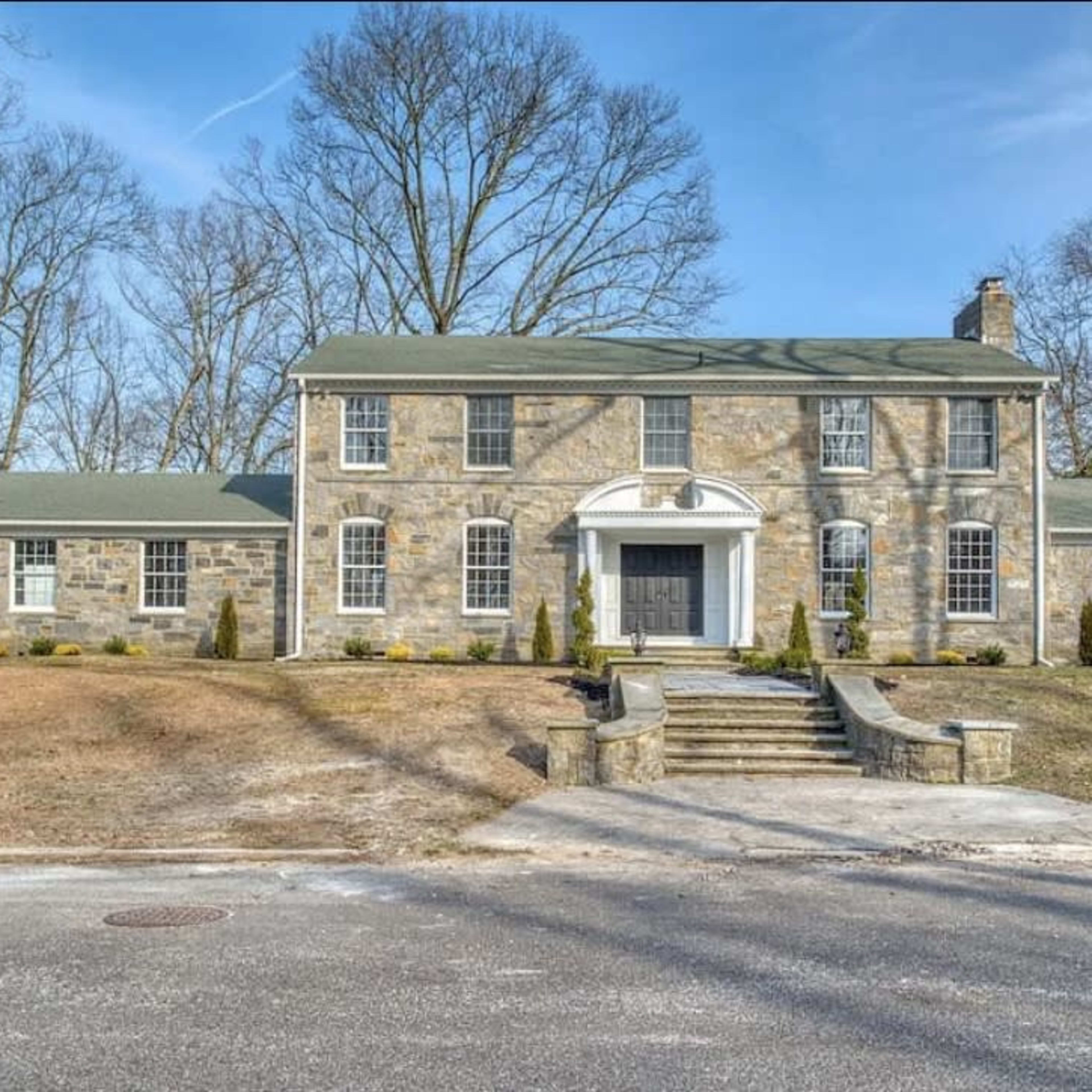 A large, two-story stone house with a symmetrical facade and a central entrance, surrounded by bare trees and a grassy lawn.