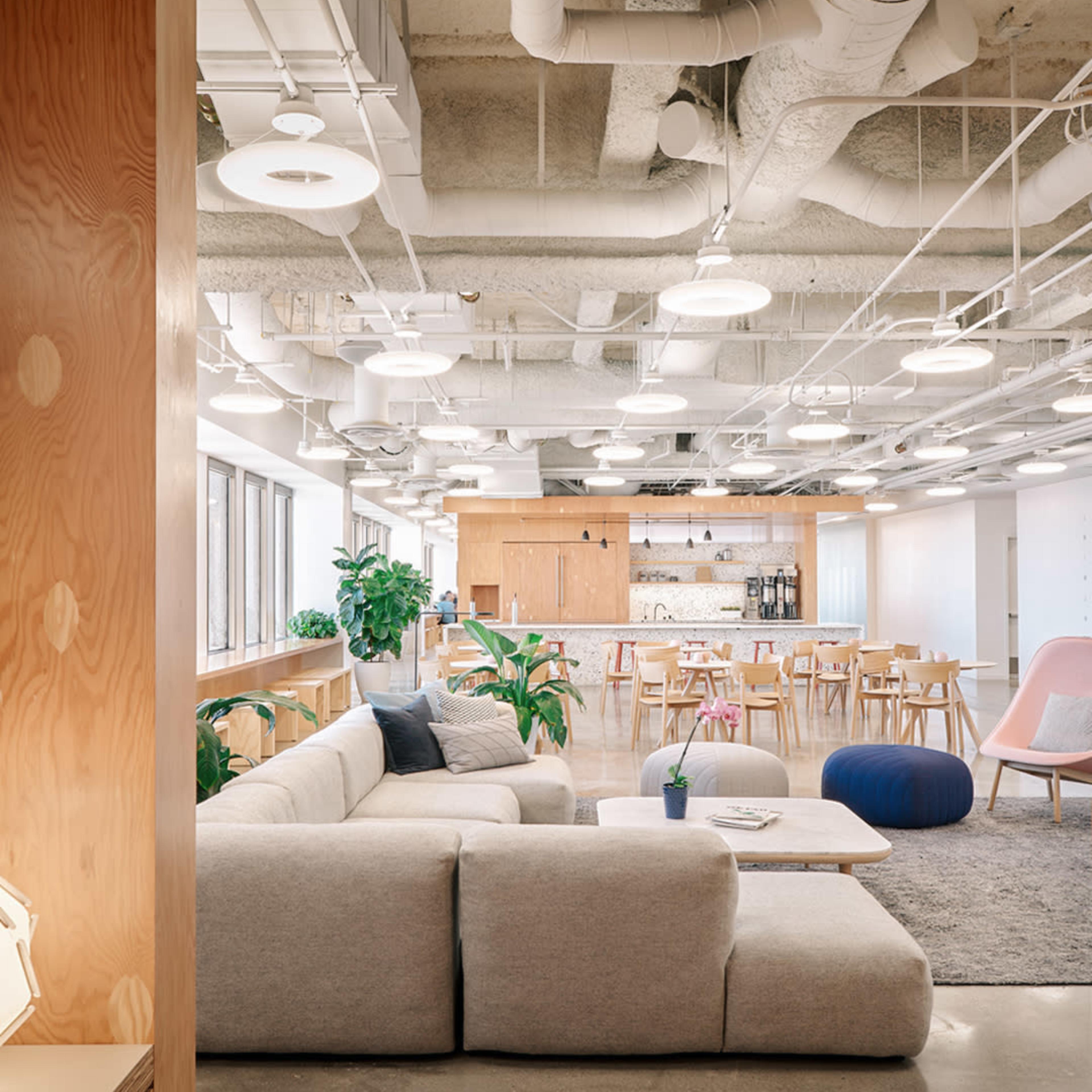 A modern office space features a cozy seating area with a light-colored sofa, plants, and a dining area with wooden furniture in the background.