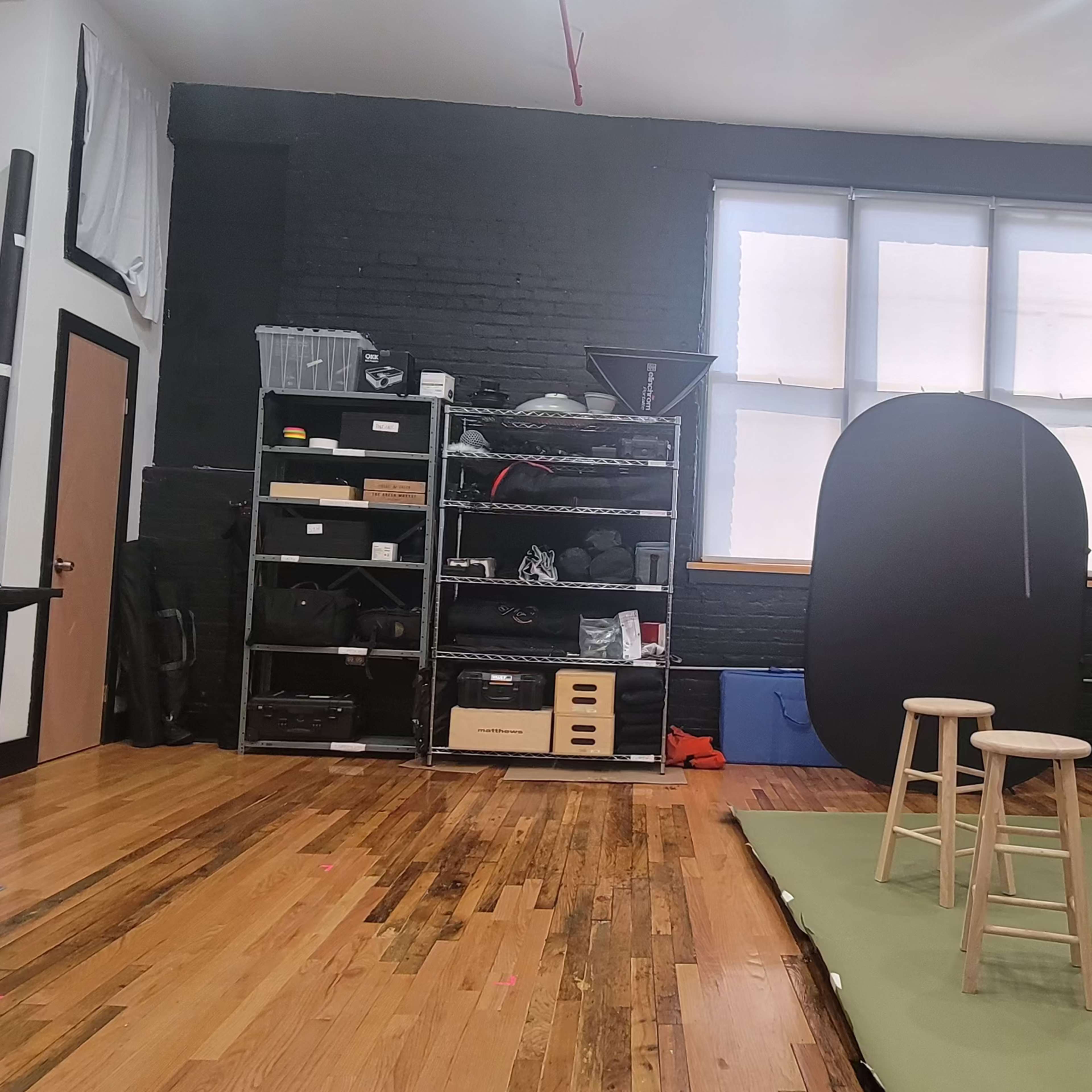 A studio space featuring a green backdrop, two wooden stools, shelving with camera equipment, and a large window.