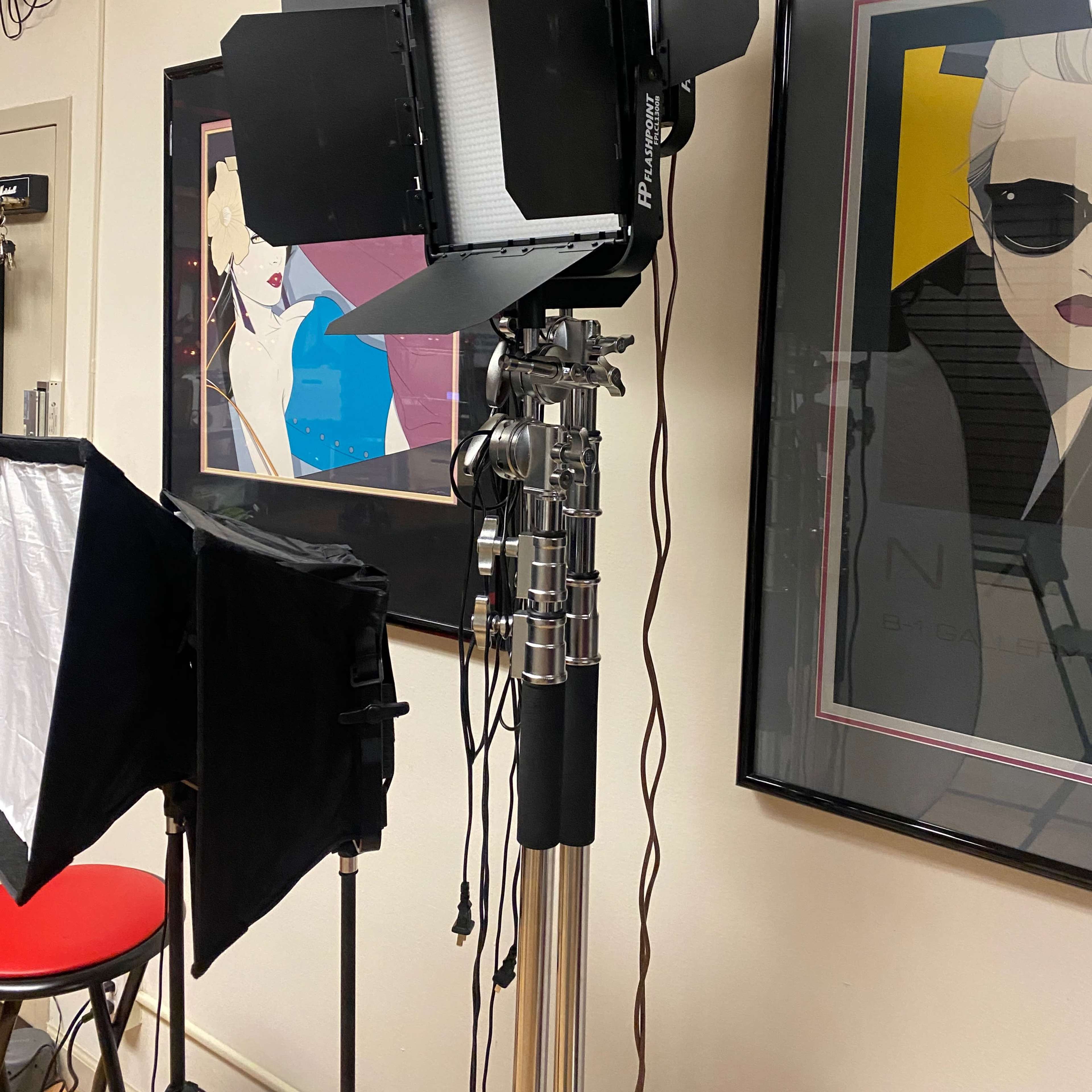 A studio lighting setup with a softbox and a floodlight mounted on a stand, positioned in front of framed artwork on a wall.