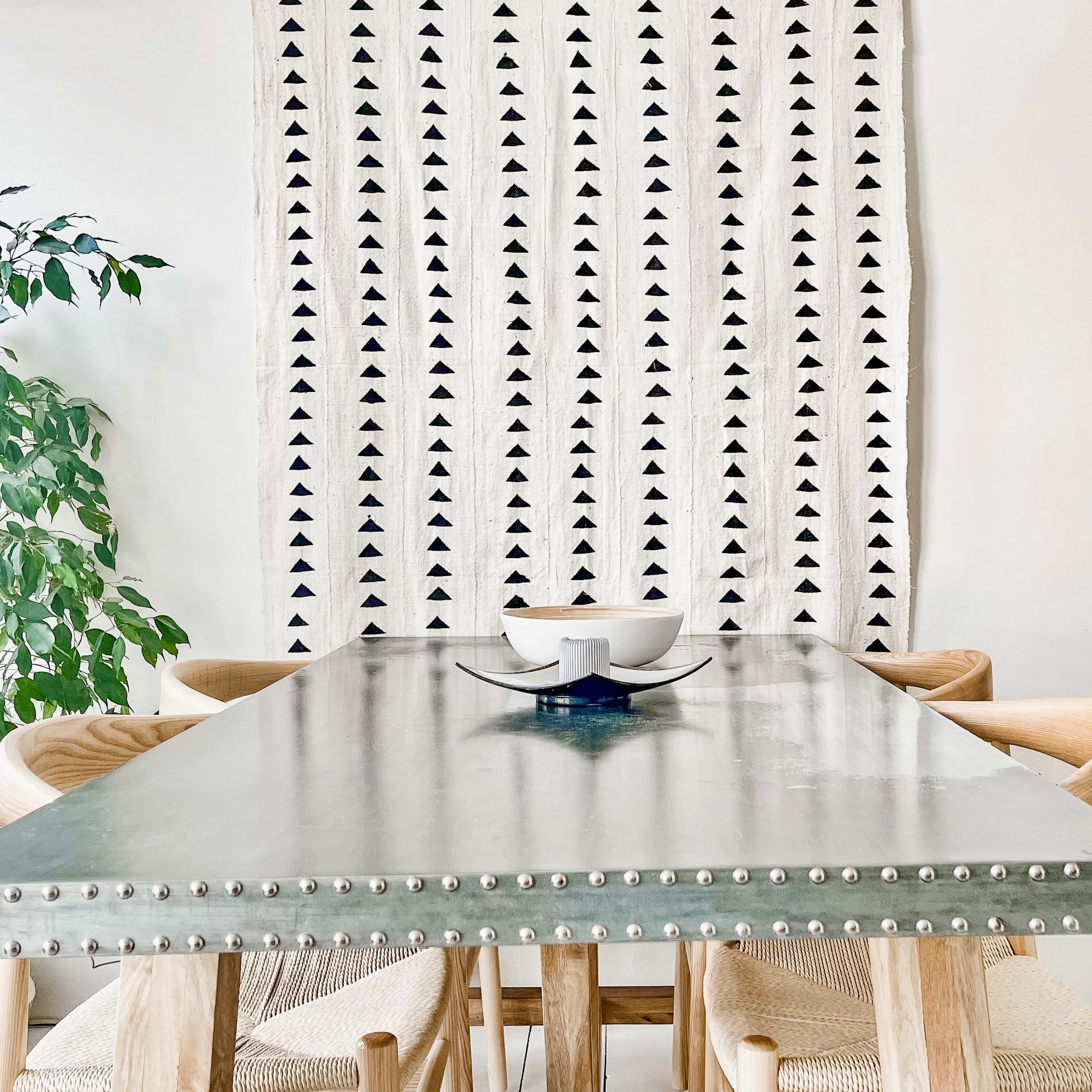 A dining table with a metal surface and studded edges is set beneath a patterned wall hanging and alongside a potted plant.