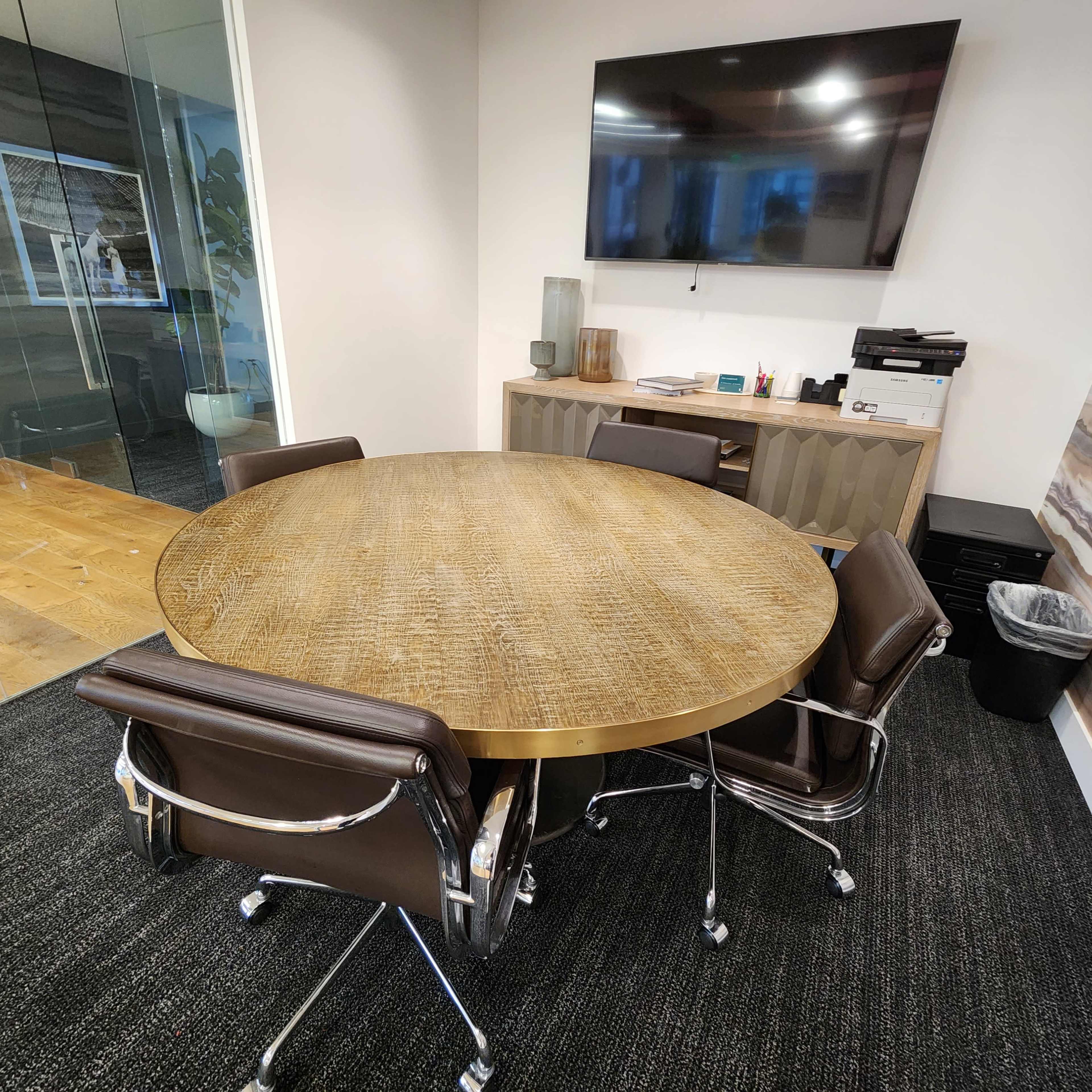 A round table with a metal surface and several brown chairs is situated in an office space with a television and a filing cabinet in the background.