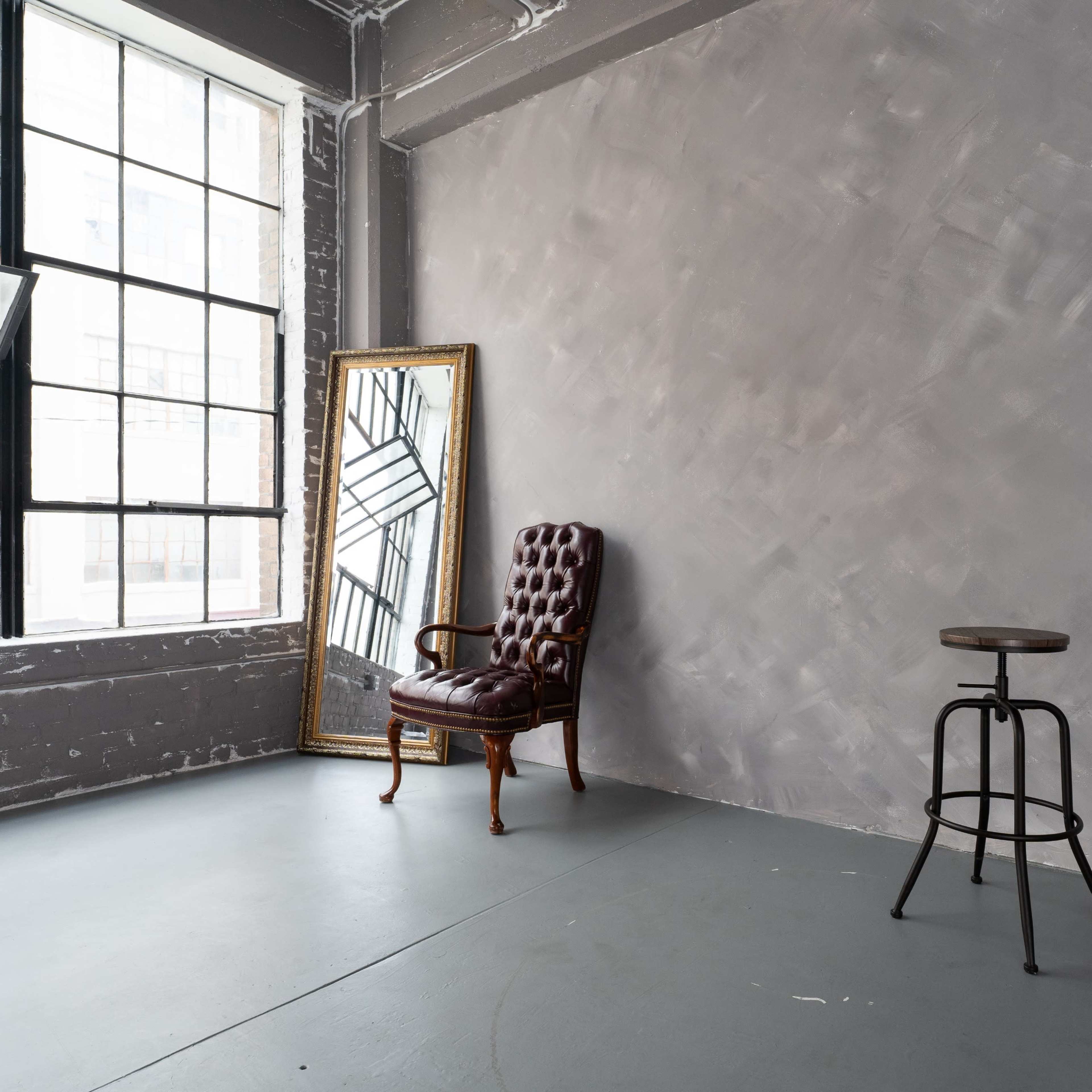 A minimalist interior with a vintage armchair and a wooden stool positioned against a gray wall, illuminated by light from large windows.