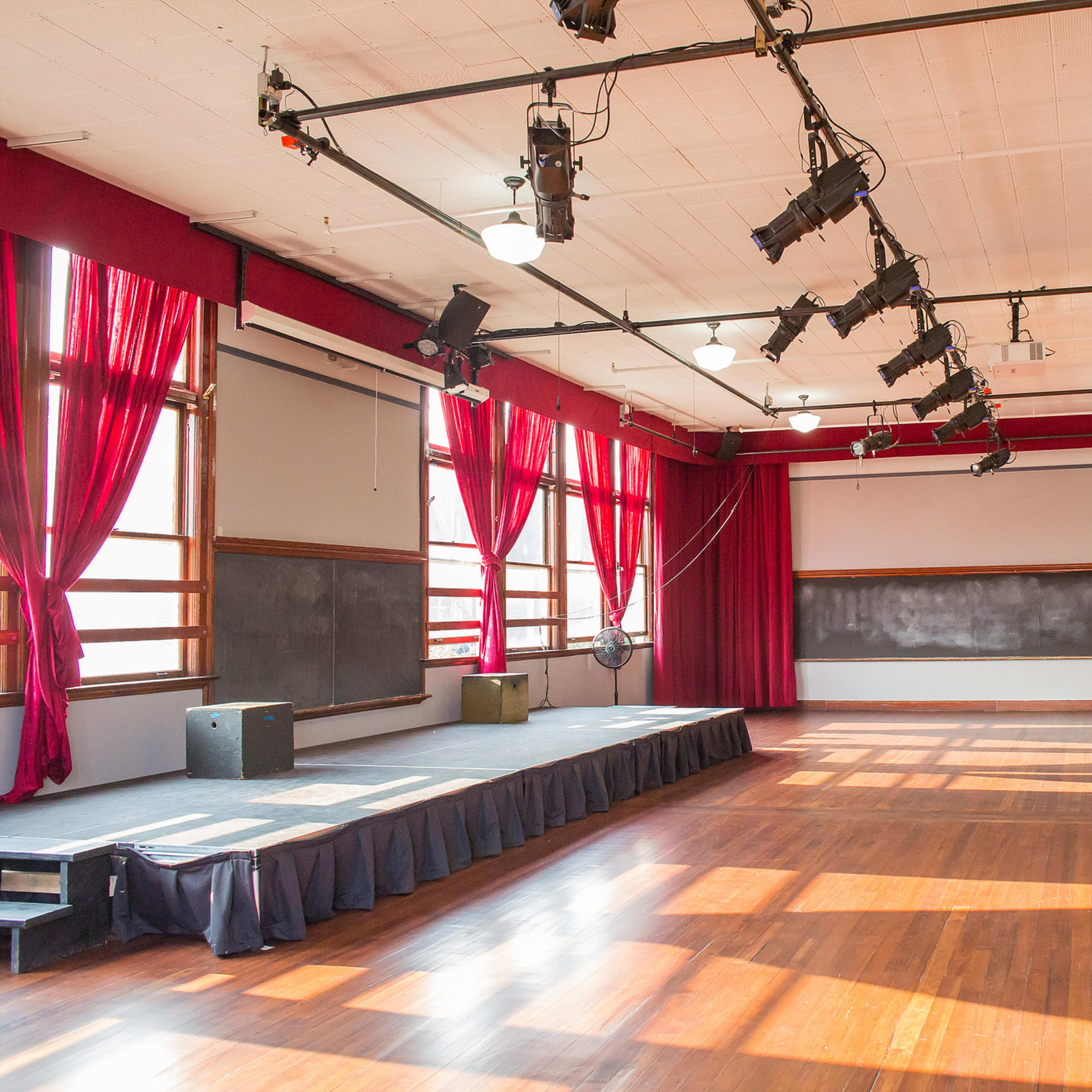 A spacious room with large windows, wooden floors, red curtains, and a stage equipped with spotlights and sound equipment.
