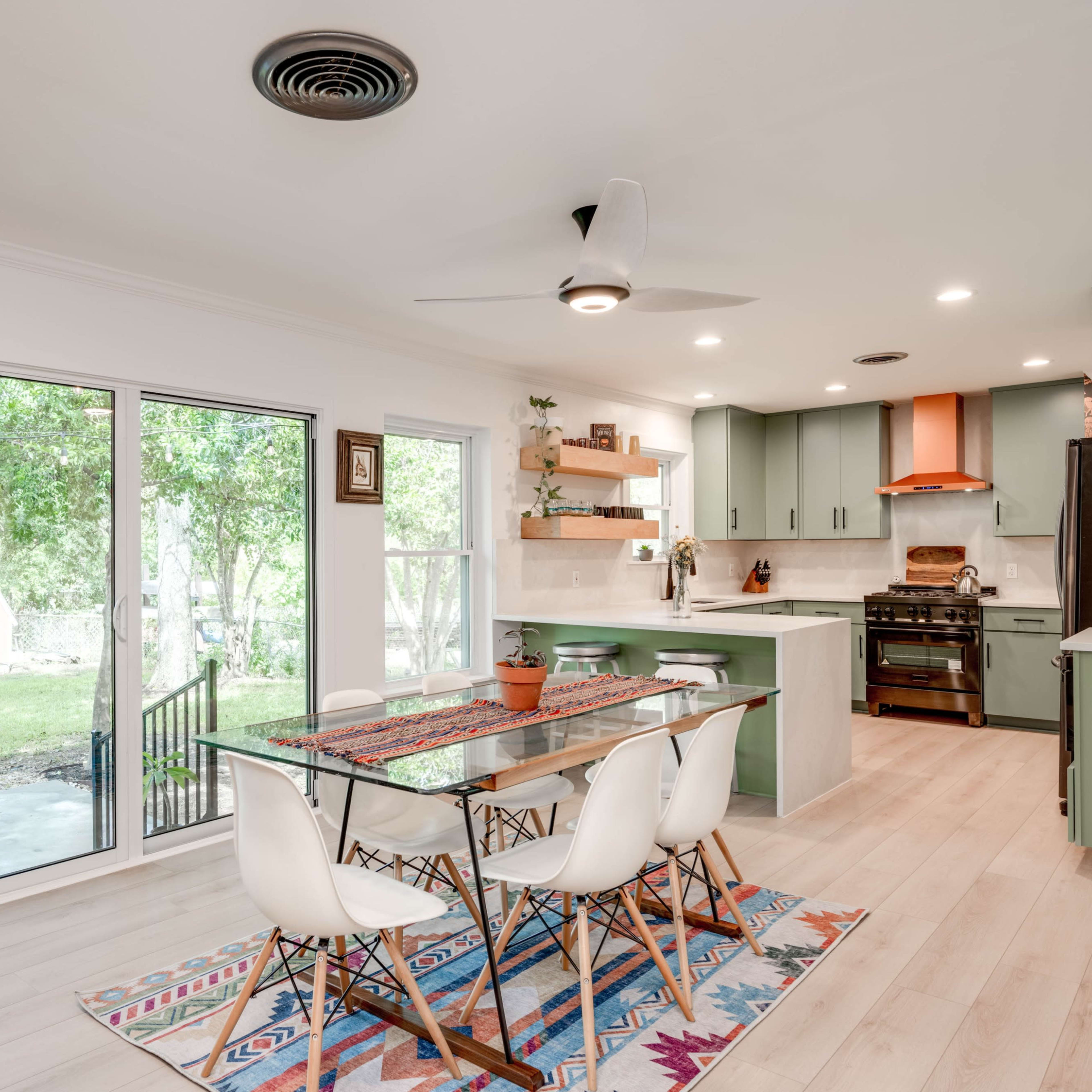 A modern kitchen and dining area features a glass table surrounded by white chairs, with large windows offering a view of the backyard.