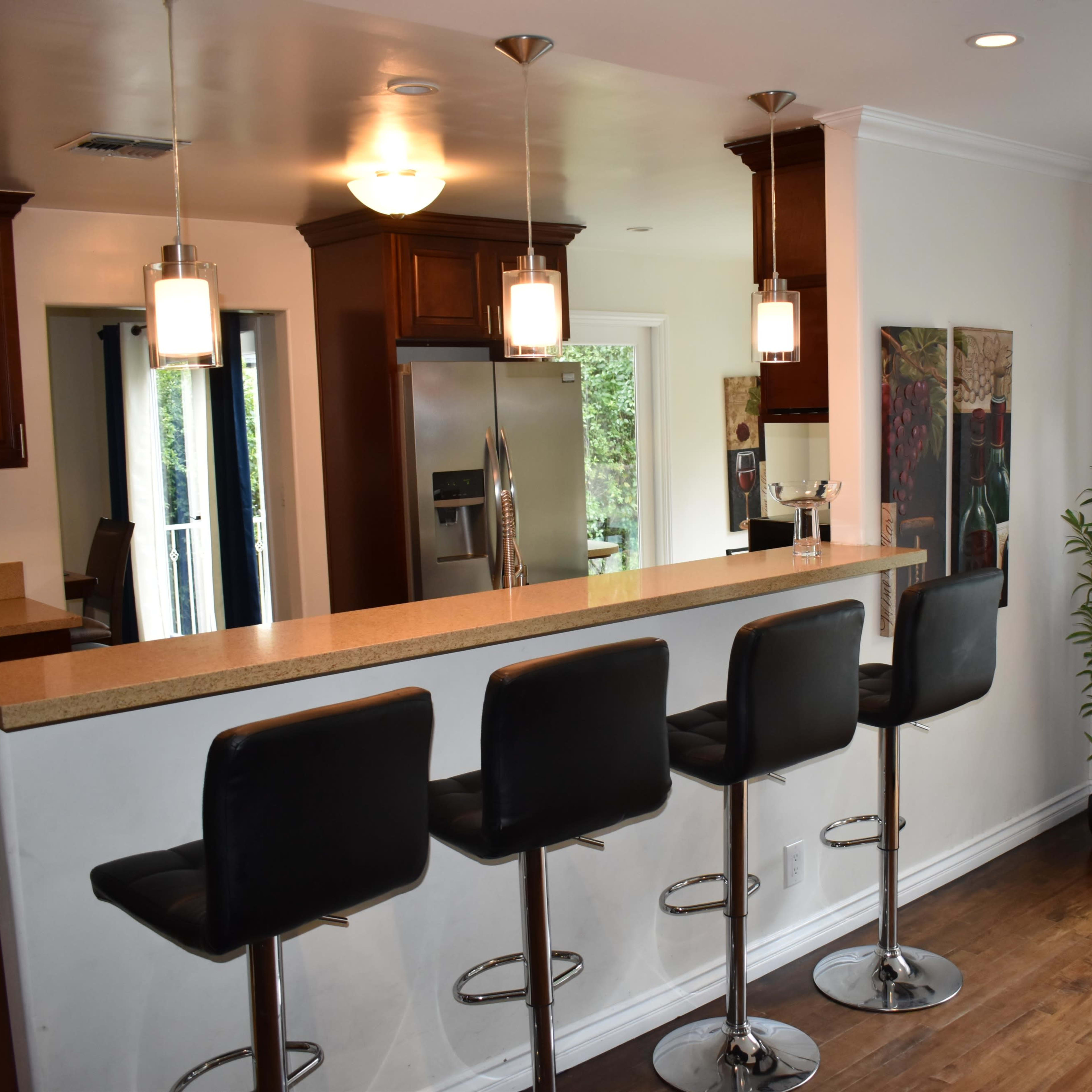 A modern kitchen with a breakfast bar featuring four black stools, pendant lighting, and a view of a garden through glass doors.
