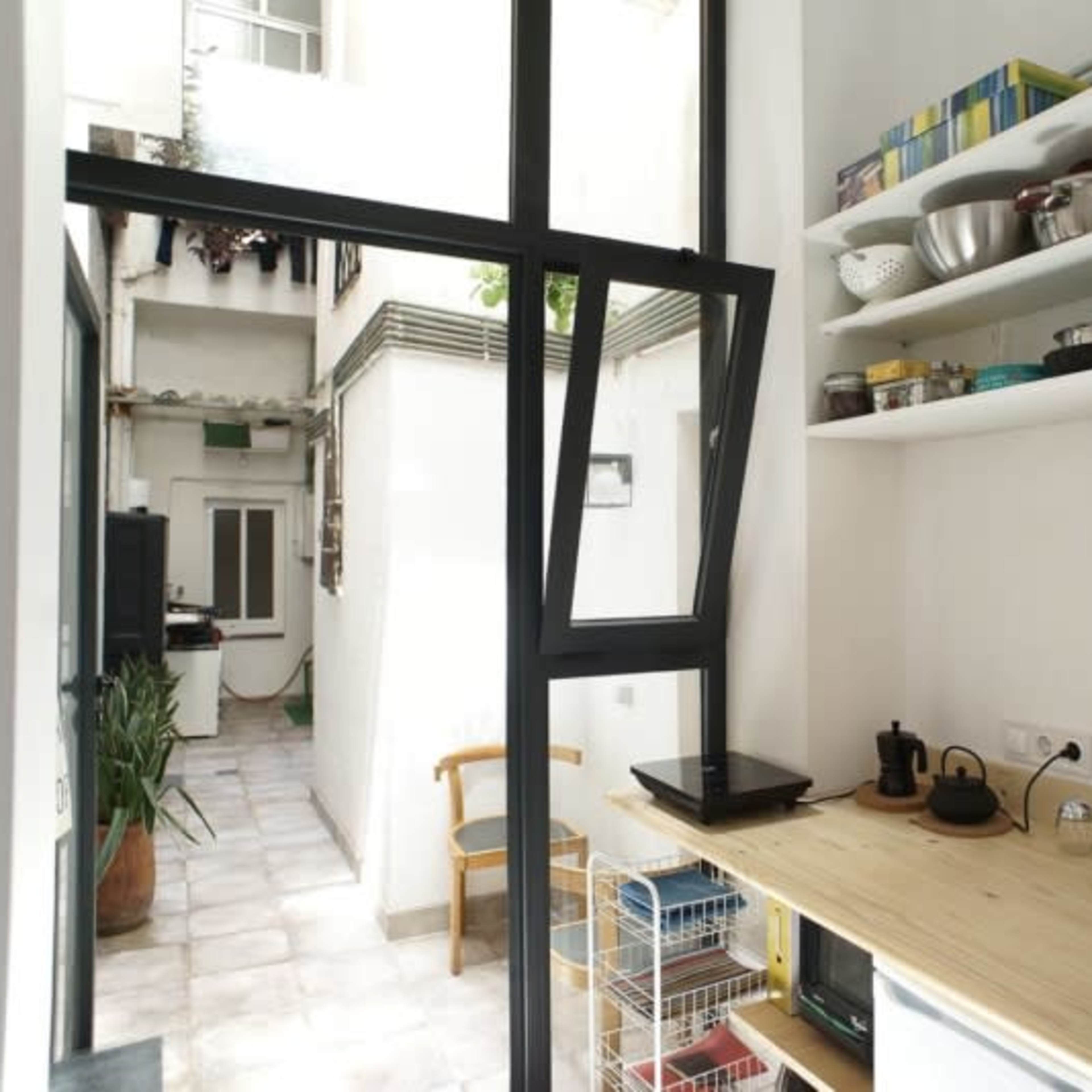 A small kitchen with a wooden countertop, shelves of dishware, and an open window overlooking a narrow courtyard.