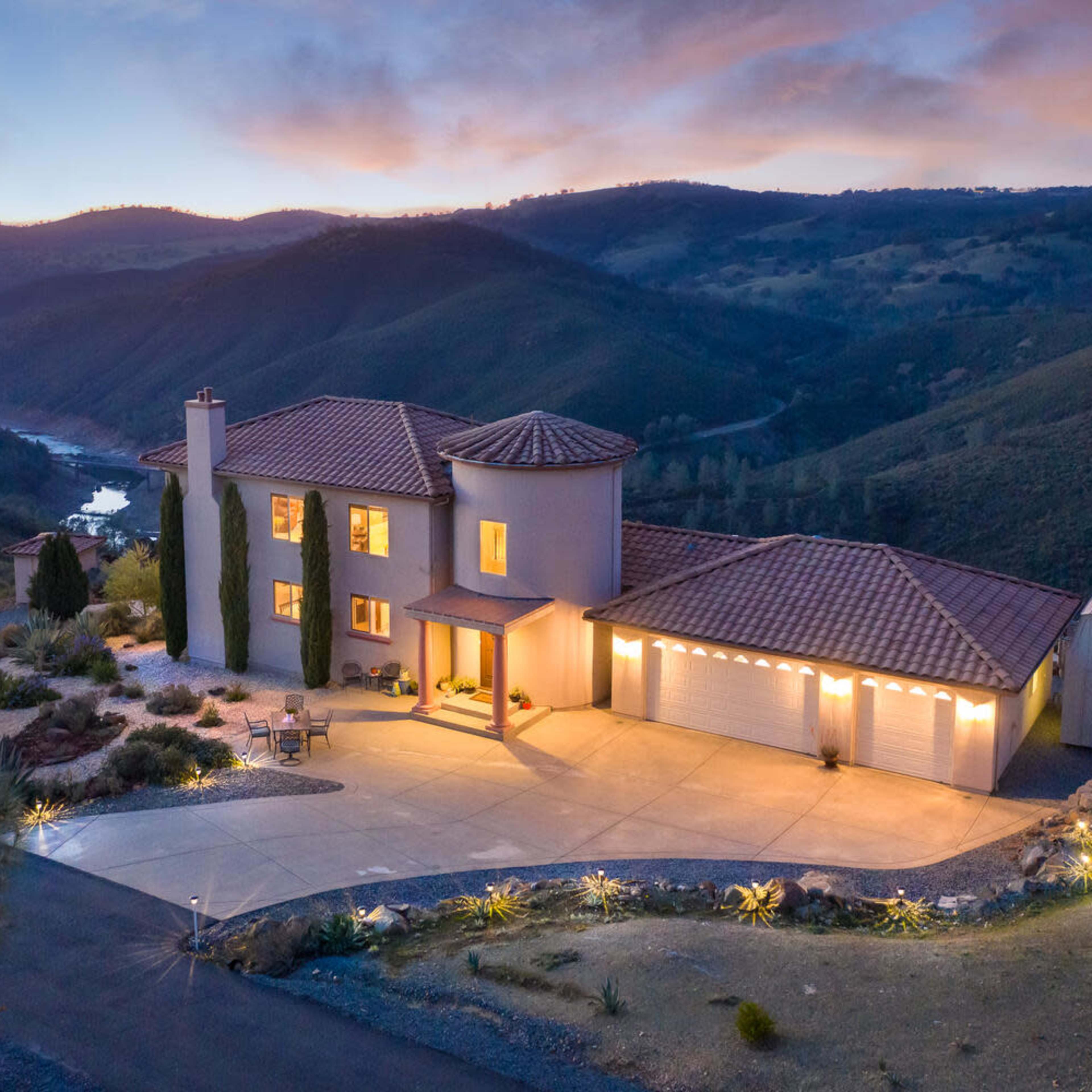 A modern two-story house with a tile roof, surrounded by rolling hills and a river, illuminated by outdoor lighting at dusk.