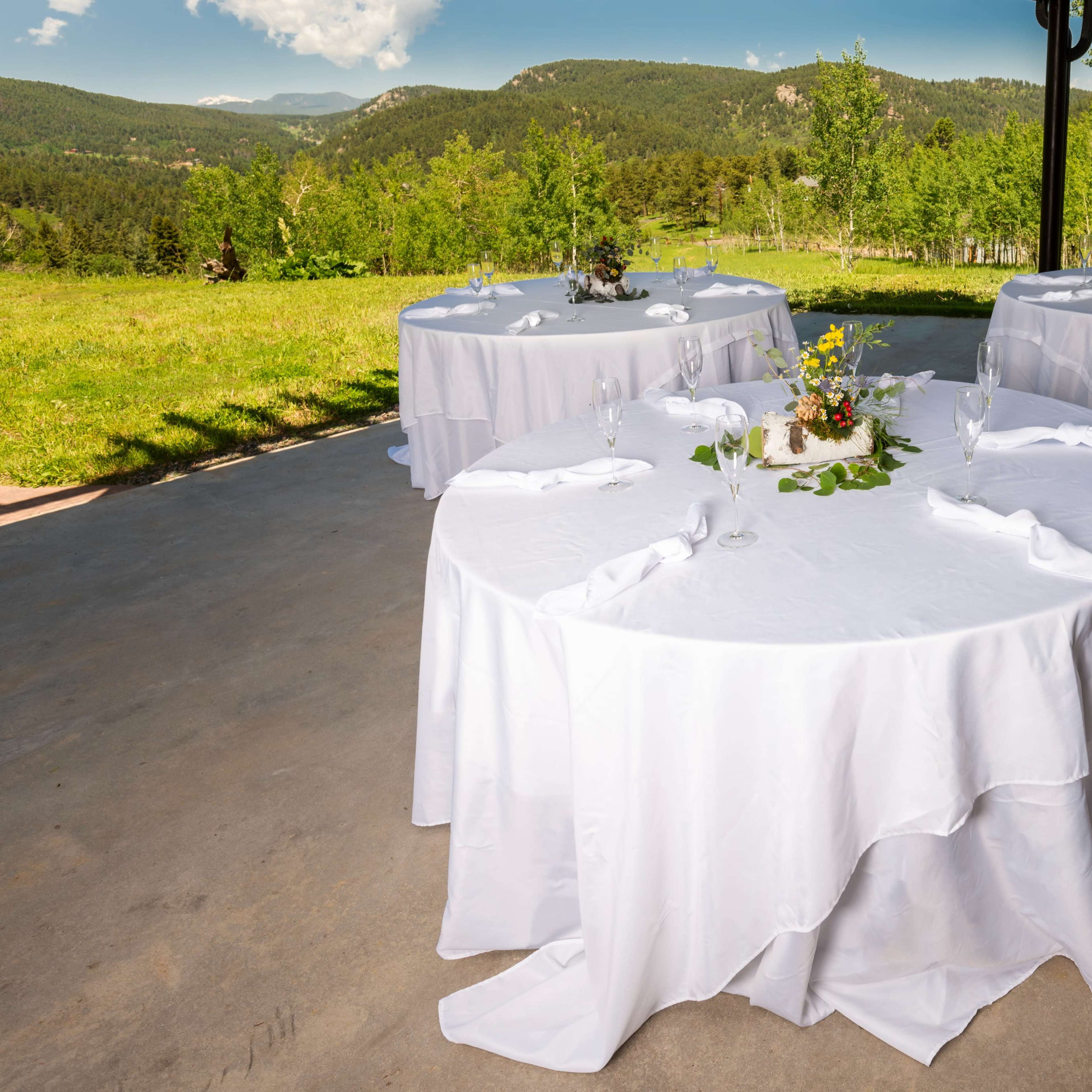 Three round tables covered with white tablecloths are arranged outdoors on a concrete surface, surrounded by lush greenery and mountains in the background.