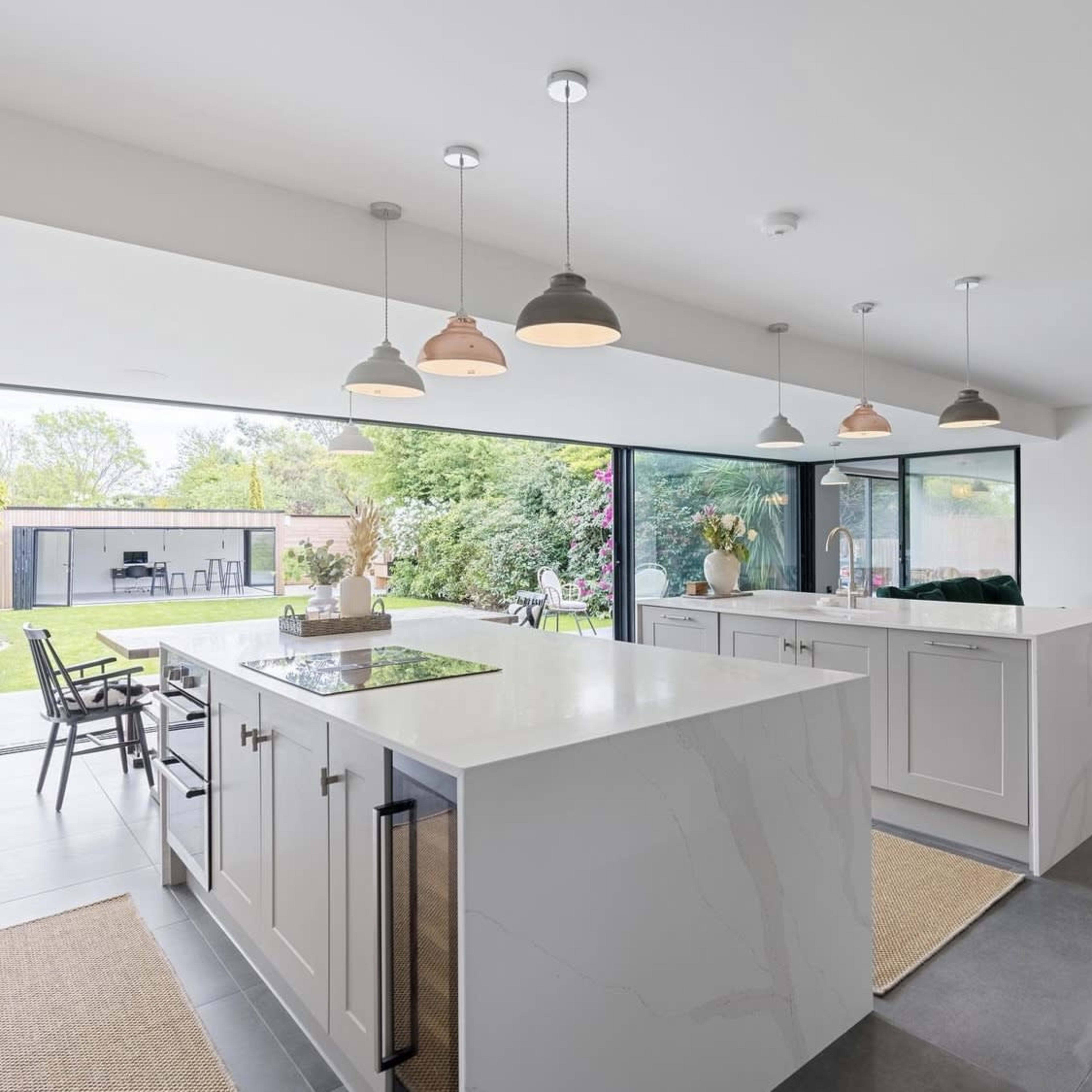 A modern kitchen features a large island with a cooktop, cabinets, and pendant lights, opening to a landscaped backyard through expansive glass doors.