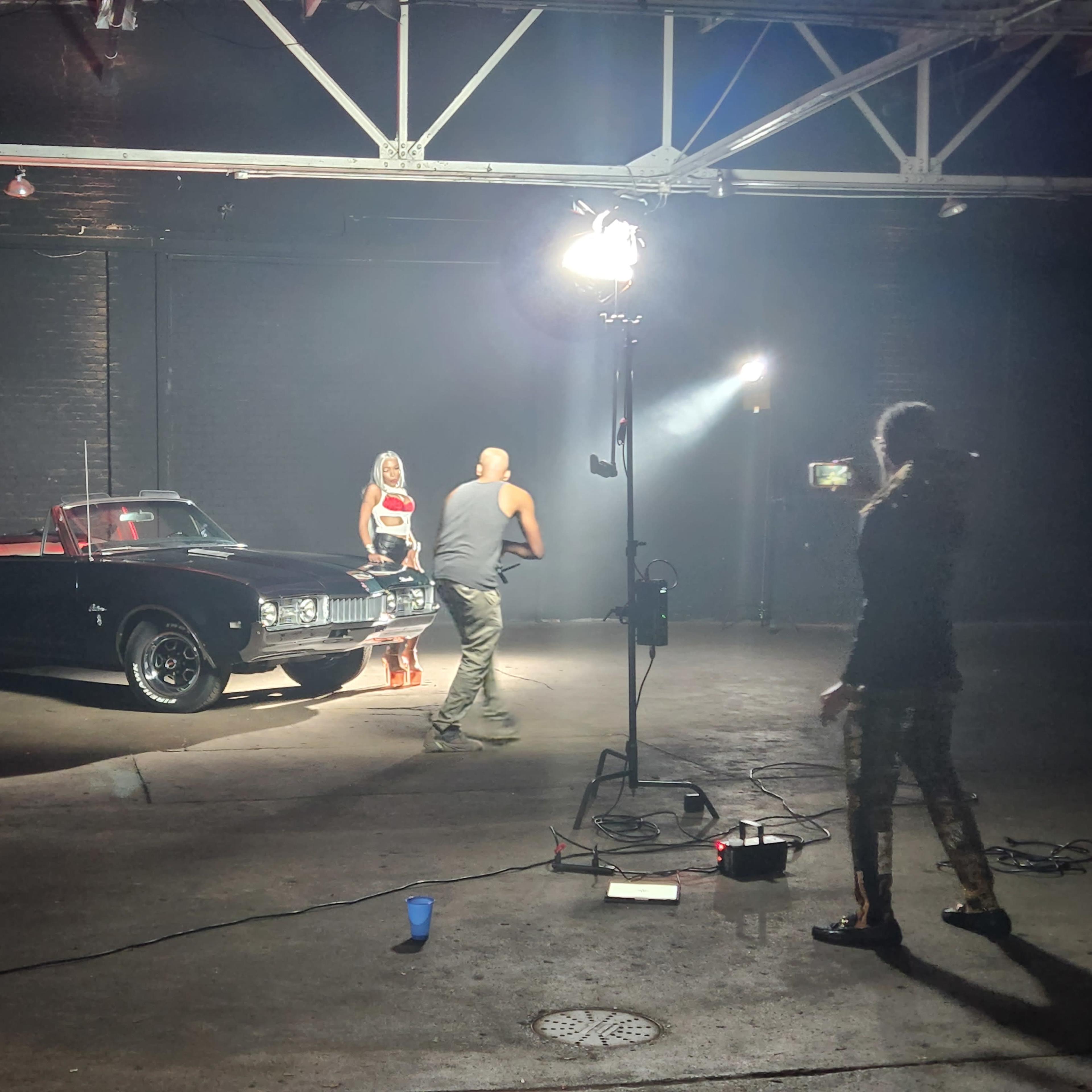 A classic black convertible car is positioned in a dimly lit industrial space while a person interacts with a model in front of it.