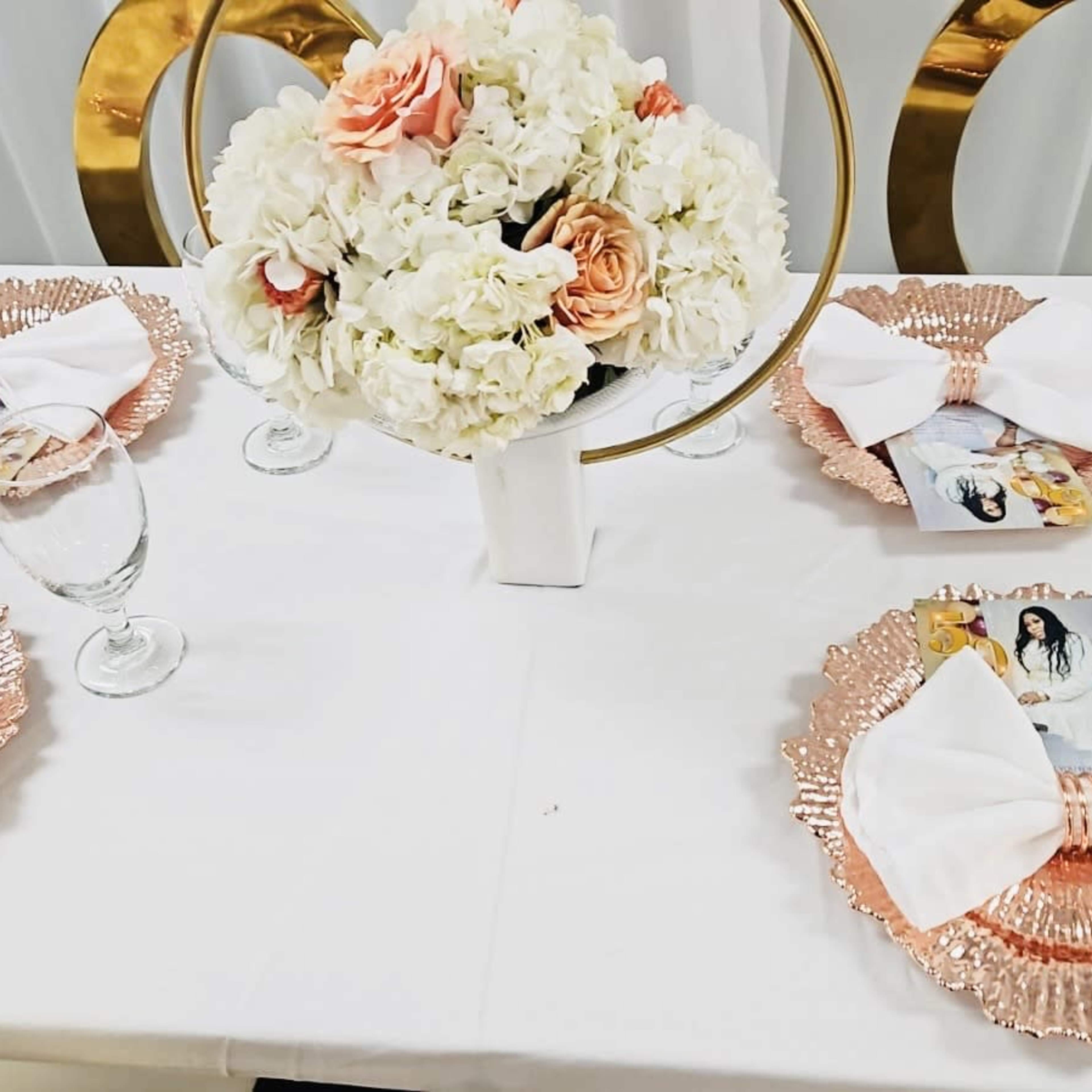A neatly arranged dining table with four places set, featuring rose gold plates and white napkins, along with a floral centerpiece.