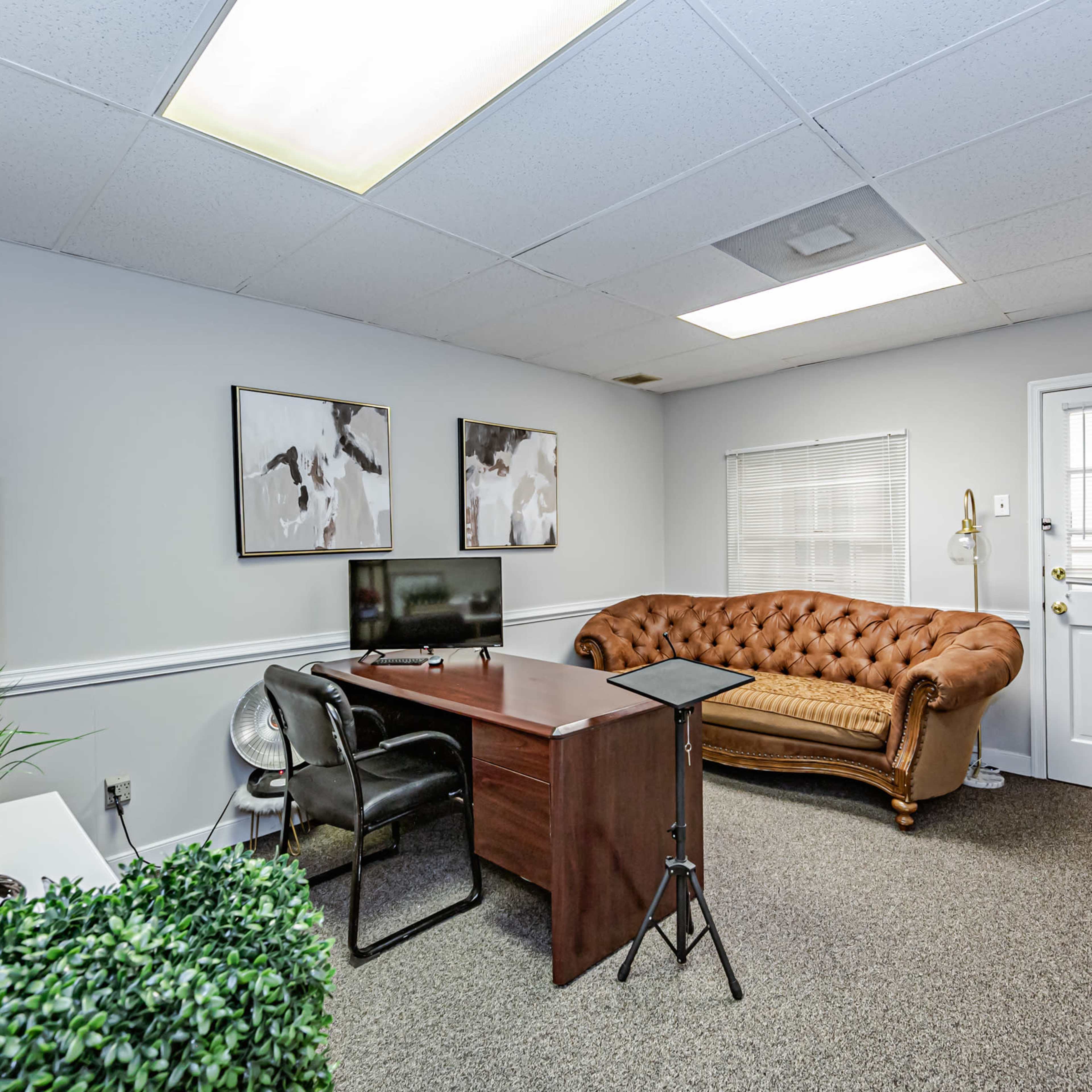 A small office space with a brown tufted couch, a wooden desk, a television, and door leading outside.