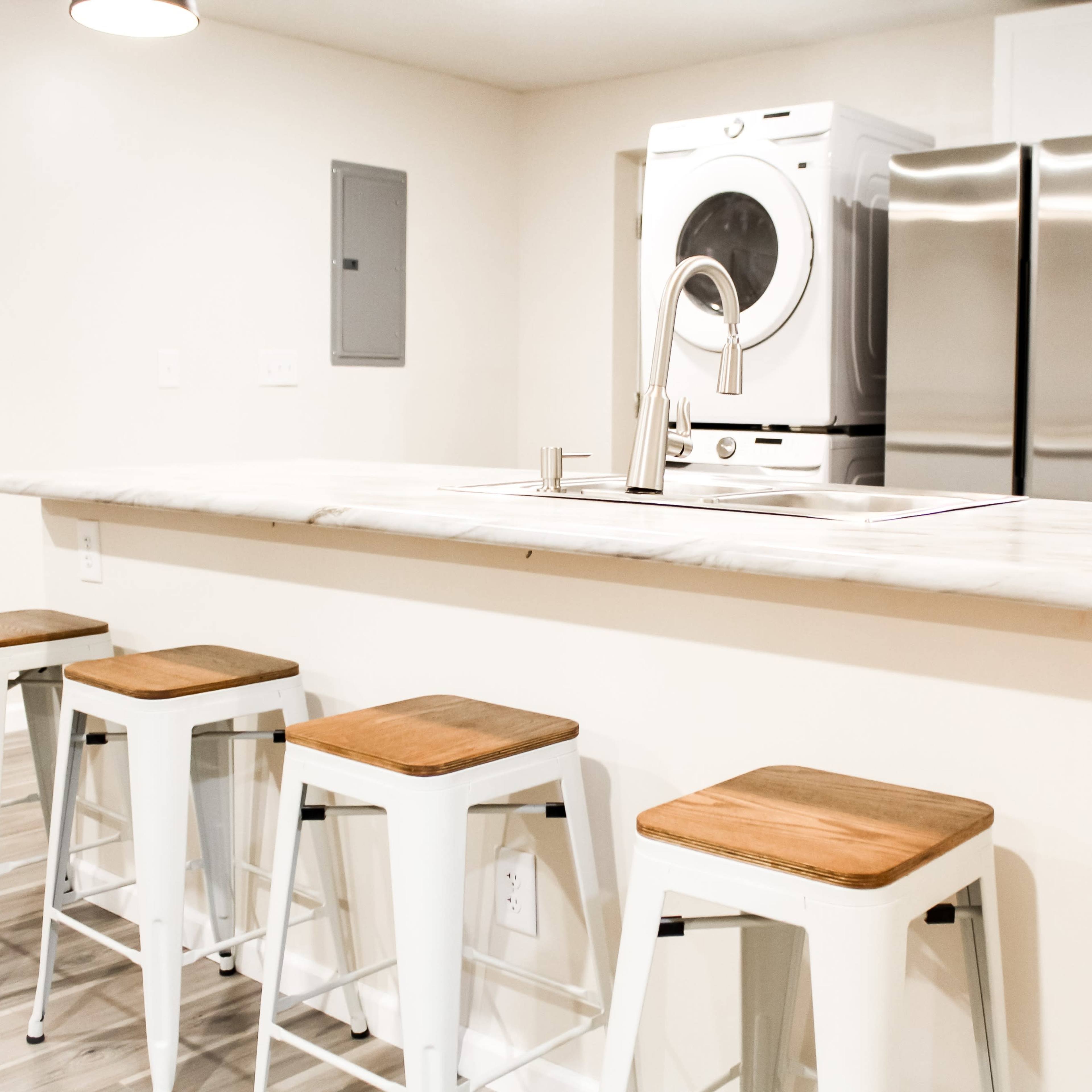 A modern kitchen with a marble-topped bar and four metal stools, featuring a washer and dryer unit and stainless steel appliances in the background.