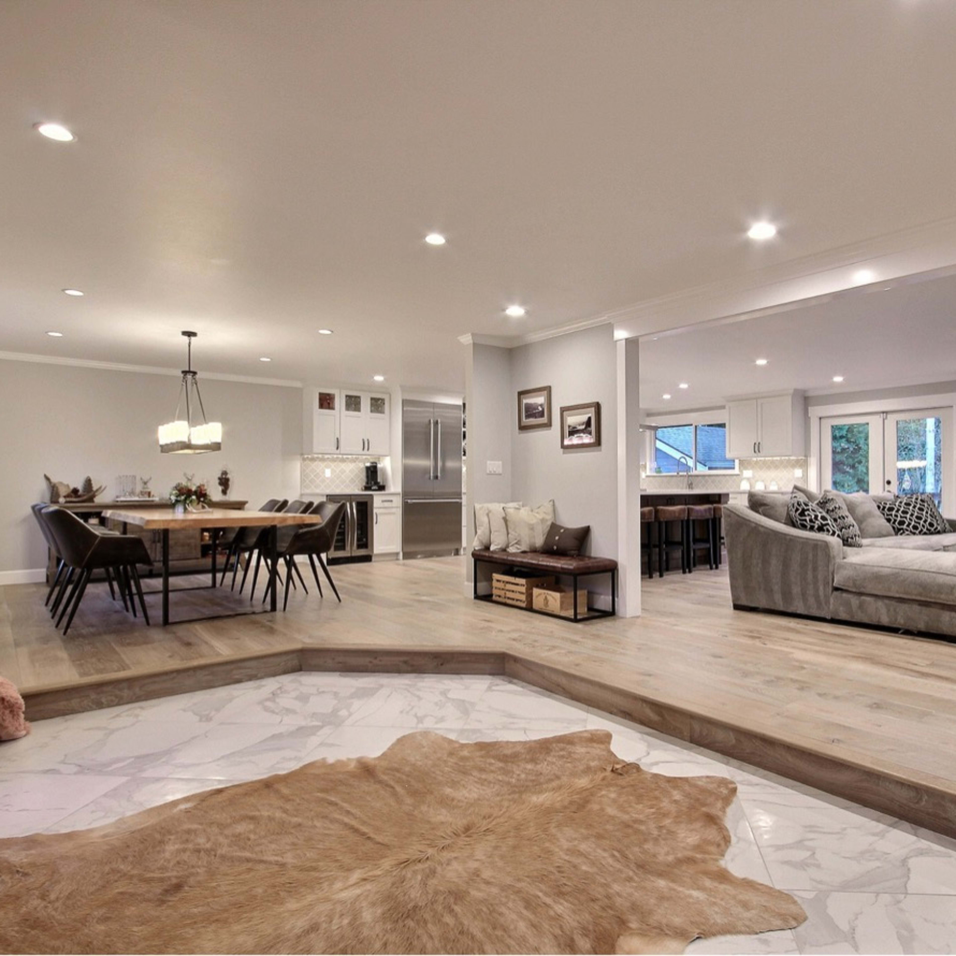 A spacious, open-concept living area features a dining space with a long table, a cozy seating area with a fireplace, and large windows allowing natural light to fill the room.